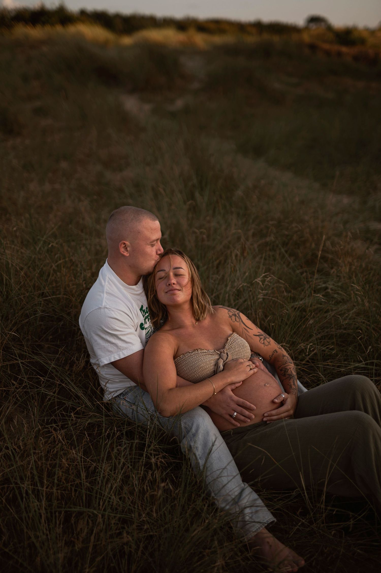 A tender maternity portrait shows a couple embracing in a field during sunset.