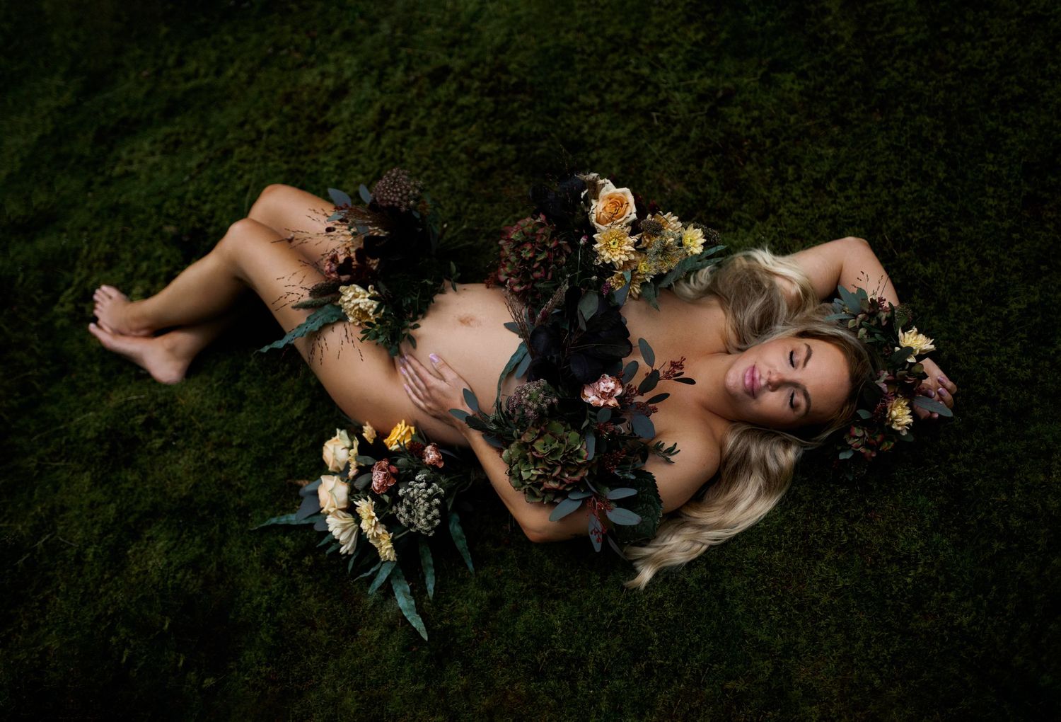 Artistic maternity photo with floral decorations on green grass in moody lighting.