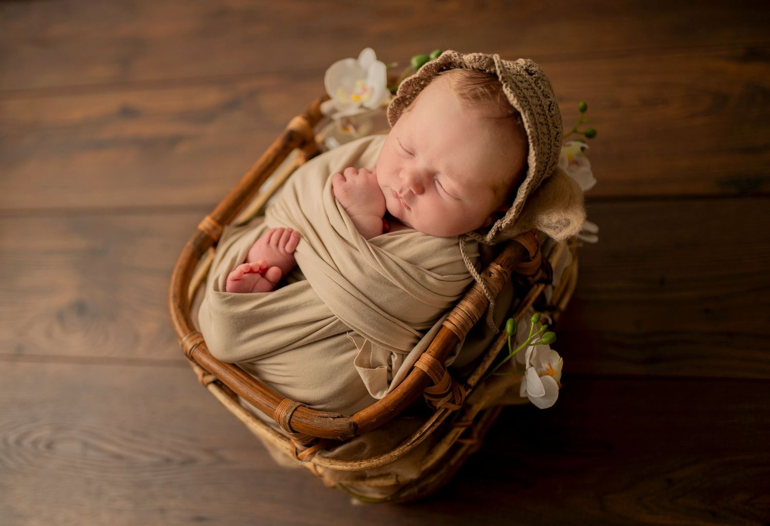 Sleeping newborn wrapped in beige fabric rests in wooden basket with white flowers.
