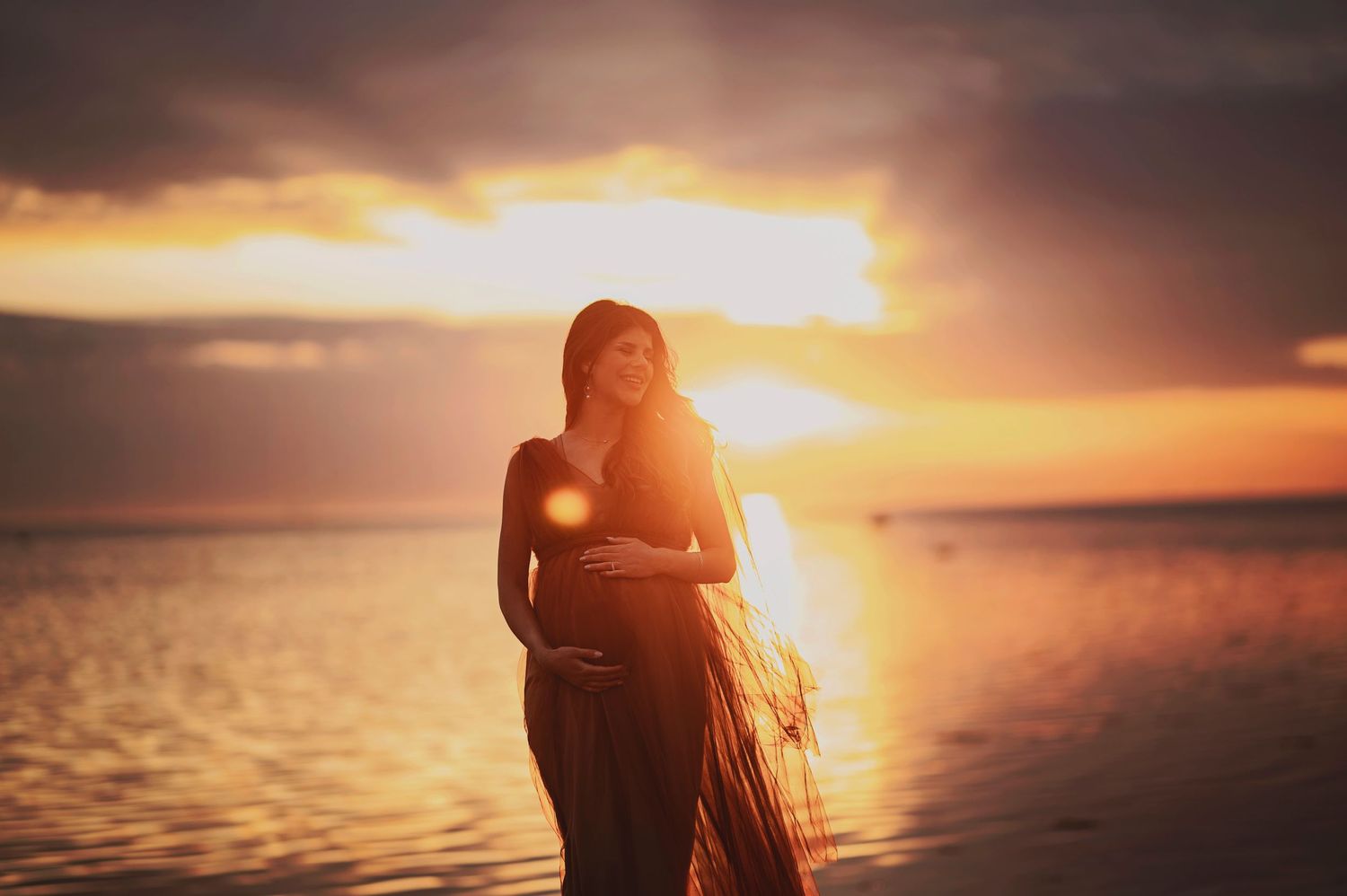 Glowing sunset creates a silhouette of an expectant mother by the ocean.