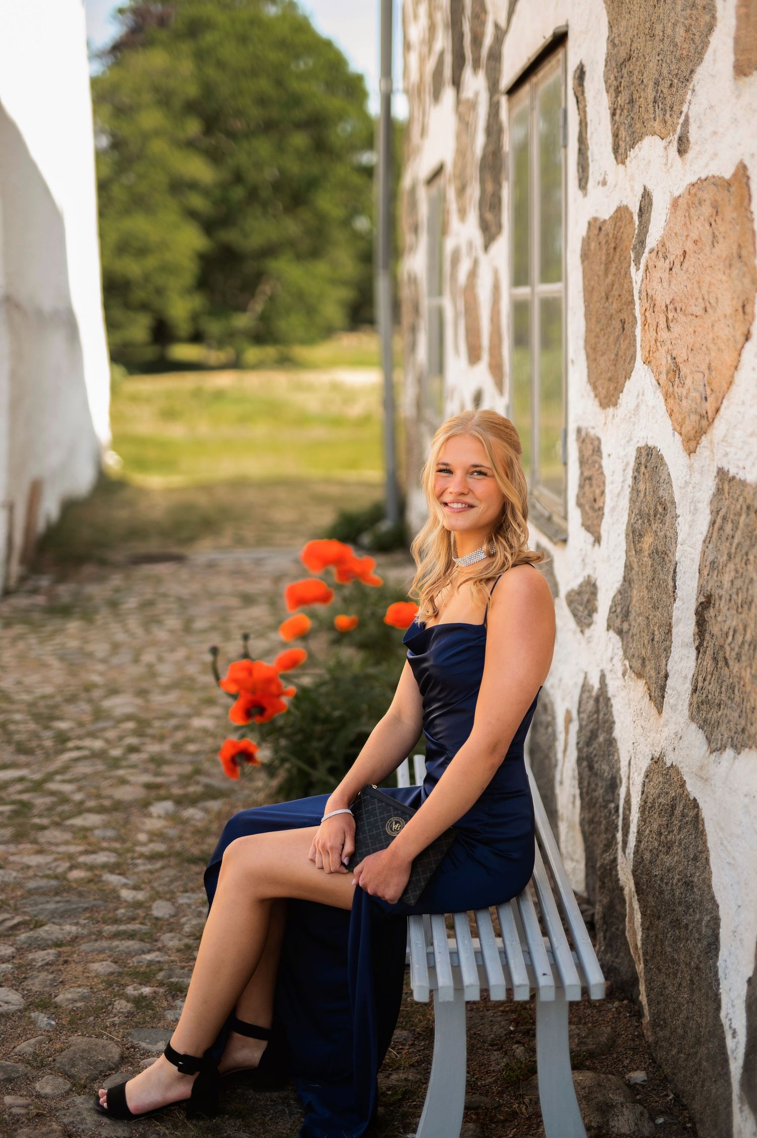 A smiling blonde woman in a navy strapless dress sits on a bench beside red poppies near a stone wall.