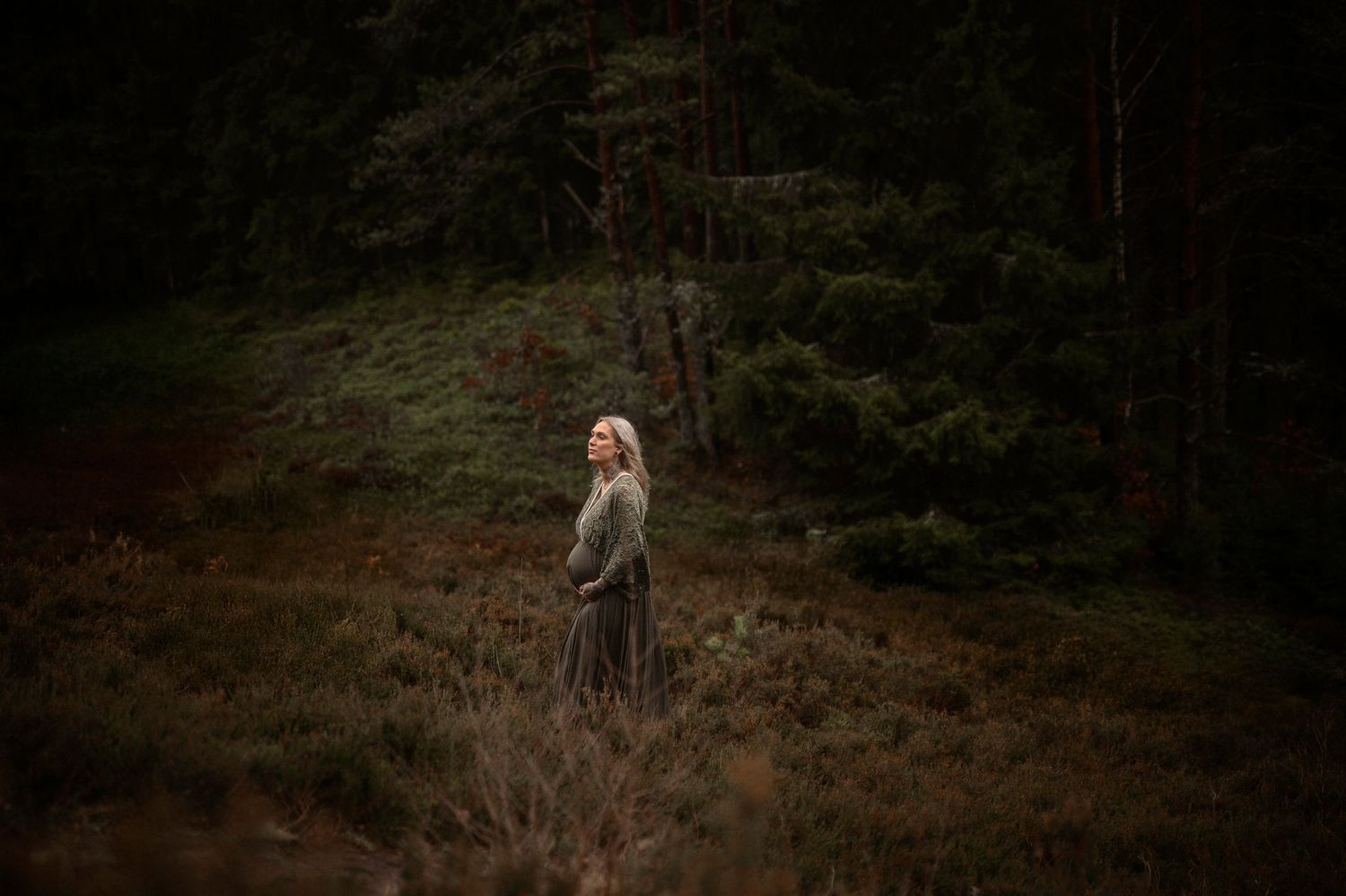 Blonde woman in a dark green forest wearing a long skirt, standing among autumn heather in a moody woodland setting.