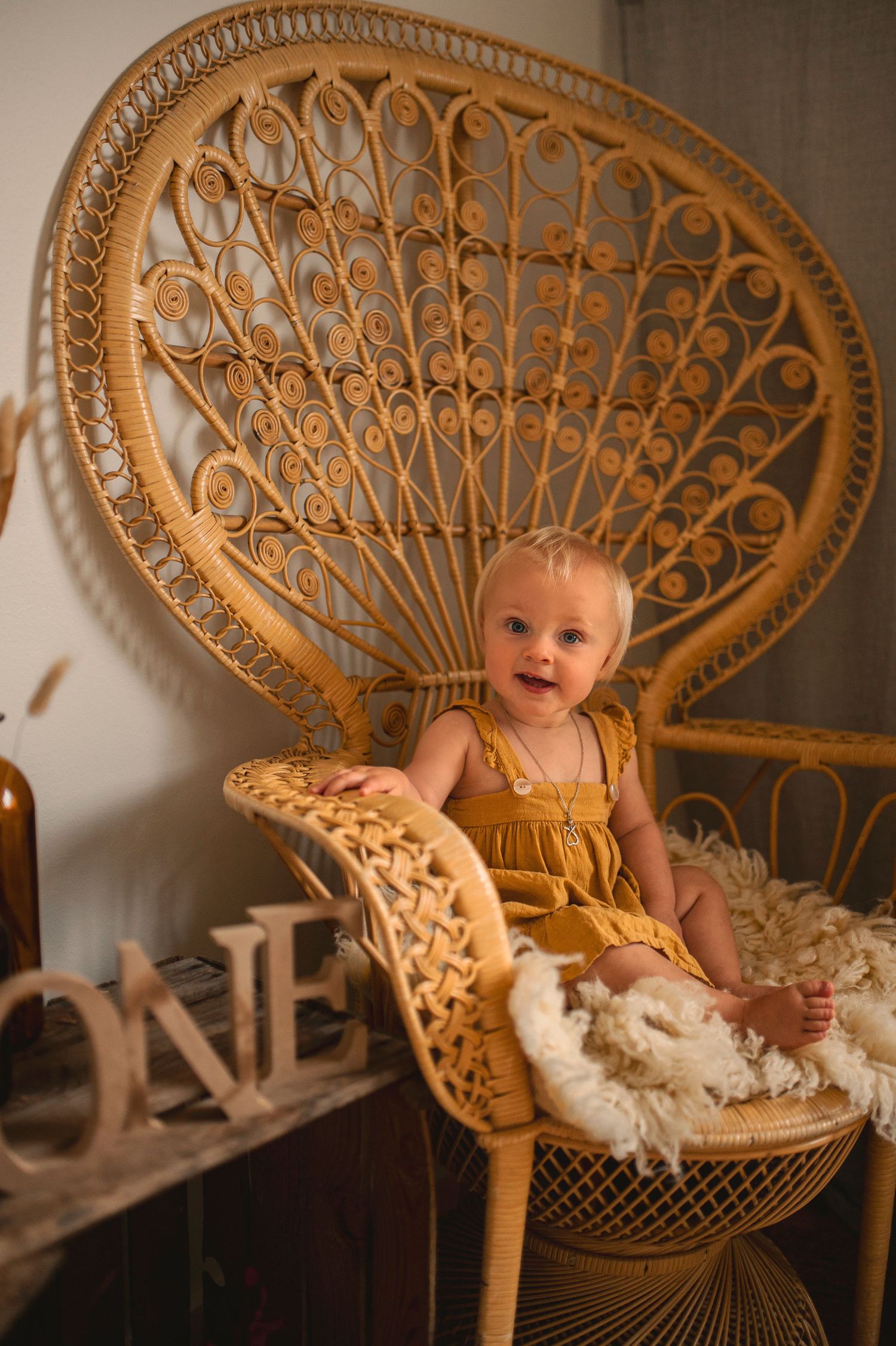 Baby in mustard romper sits happily in large wicker peacock chair with cream sheepskin.