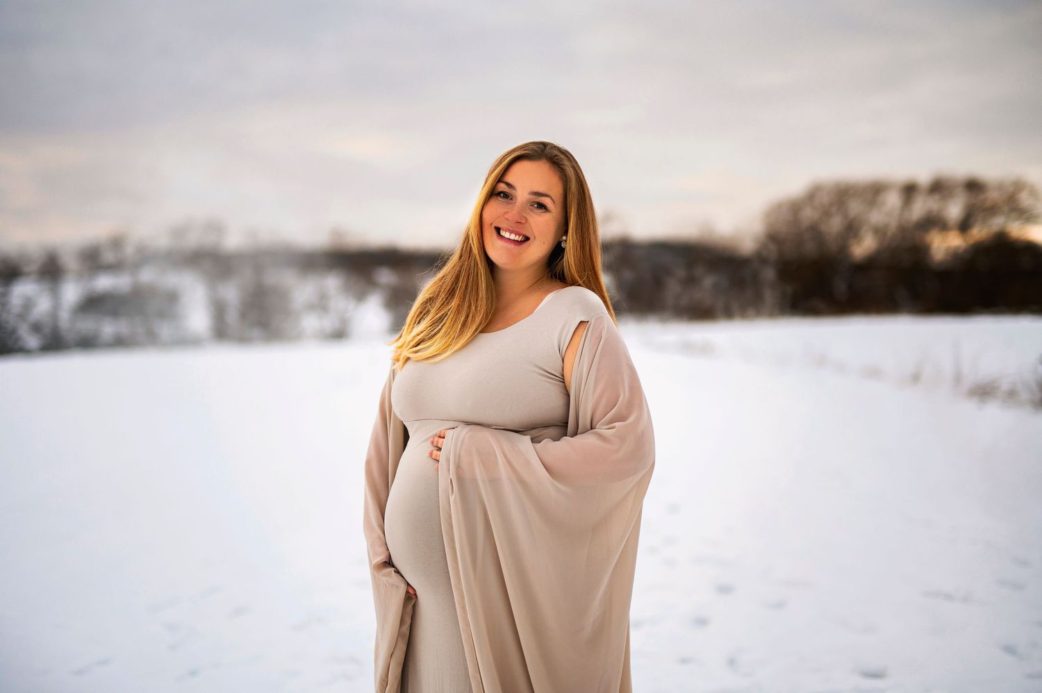Expectant mother in a flowing beige dress poses in a snowy winter landscape with a bright smile.
