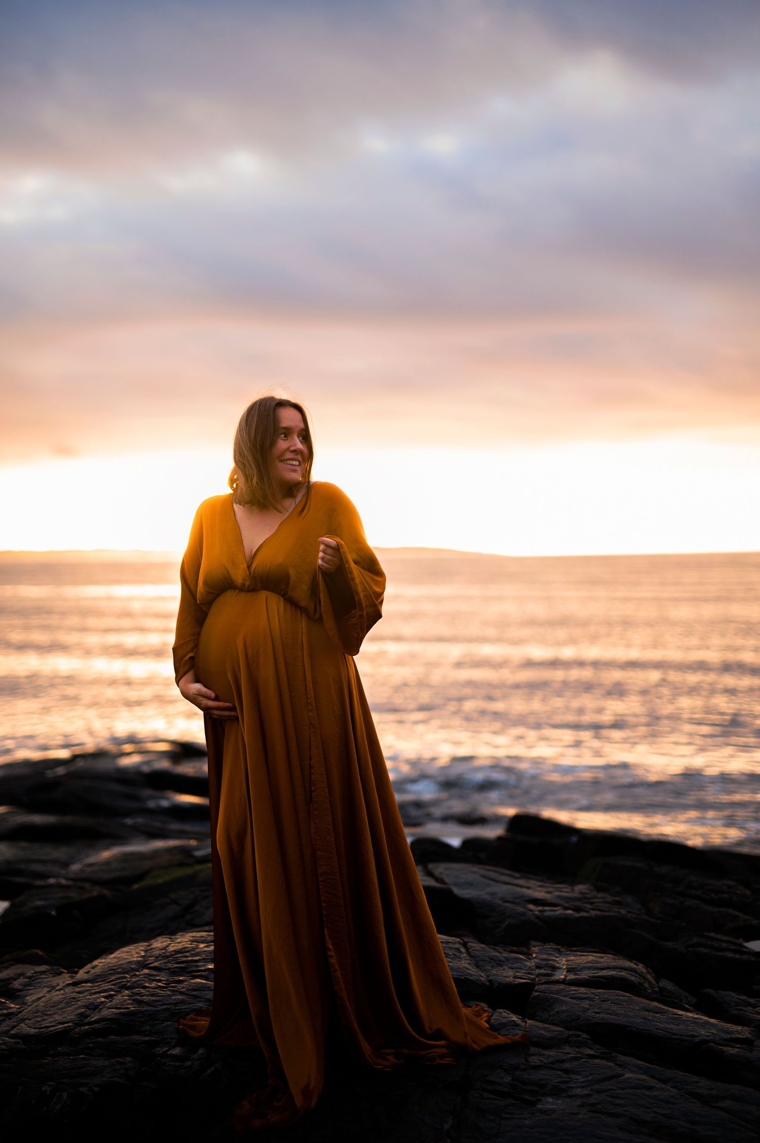 Majestic sunset maternity portrait on rocky coastline with figure in flowing mustard gown.
