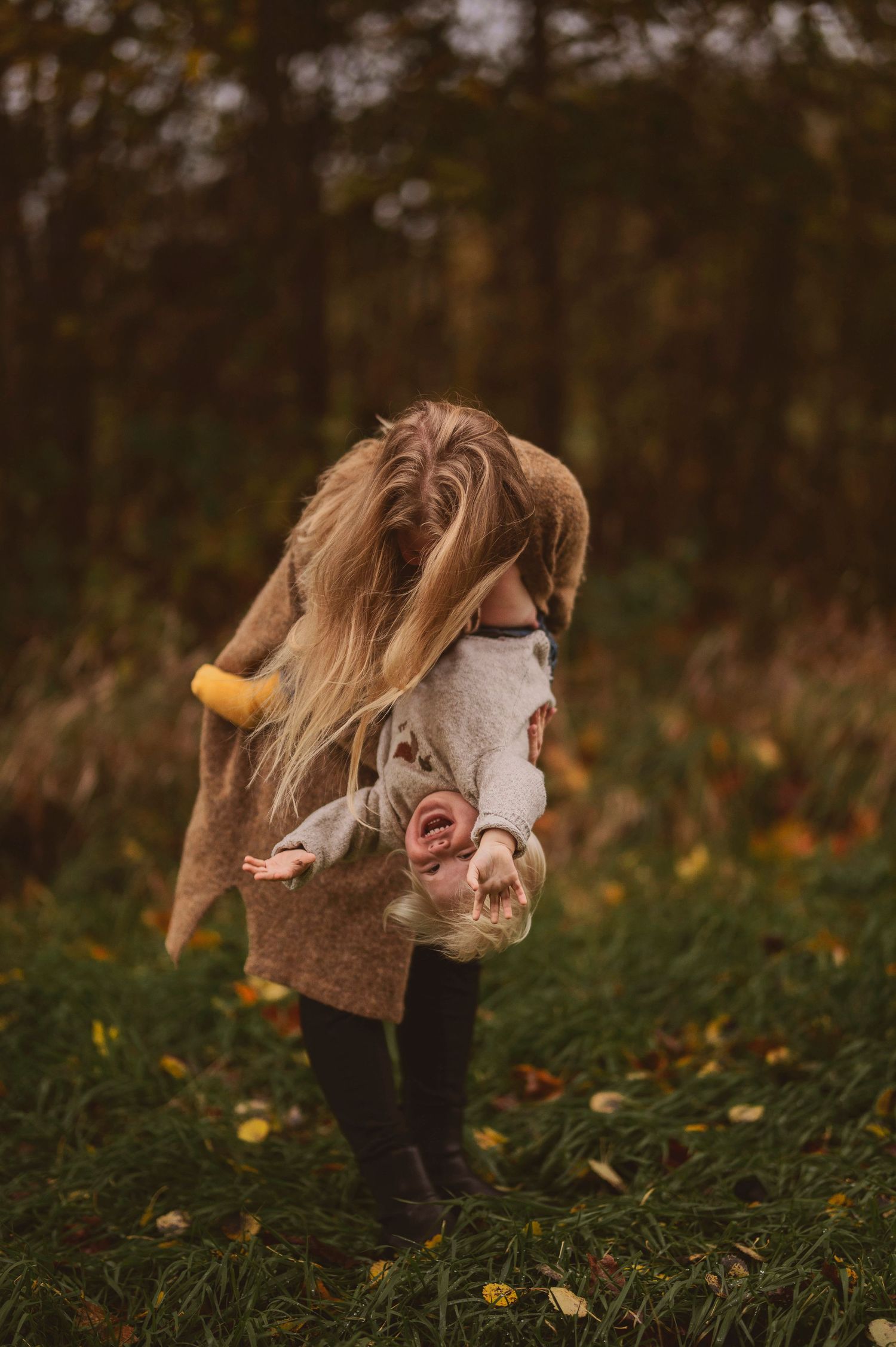 Figure in brown coat playing with happy child wearing yellow socks among autumn leaves.
