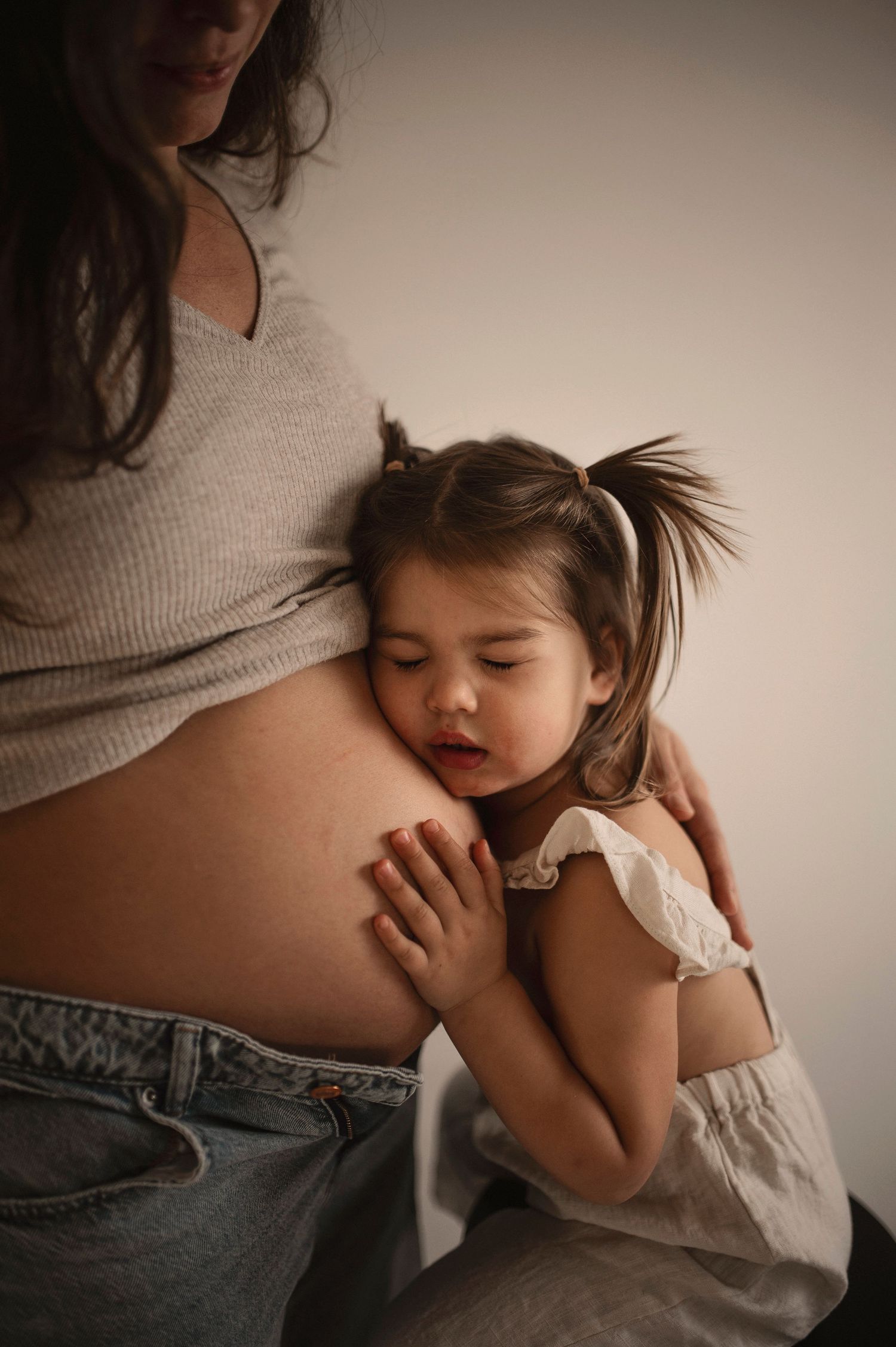 Young child lovingly embraces pregnant belly while wearing white dress.