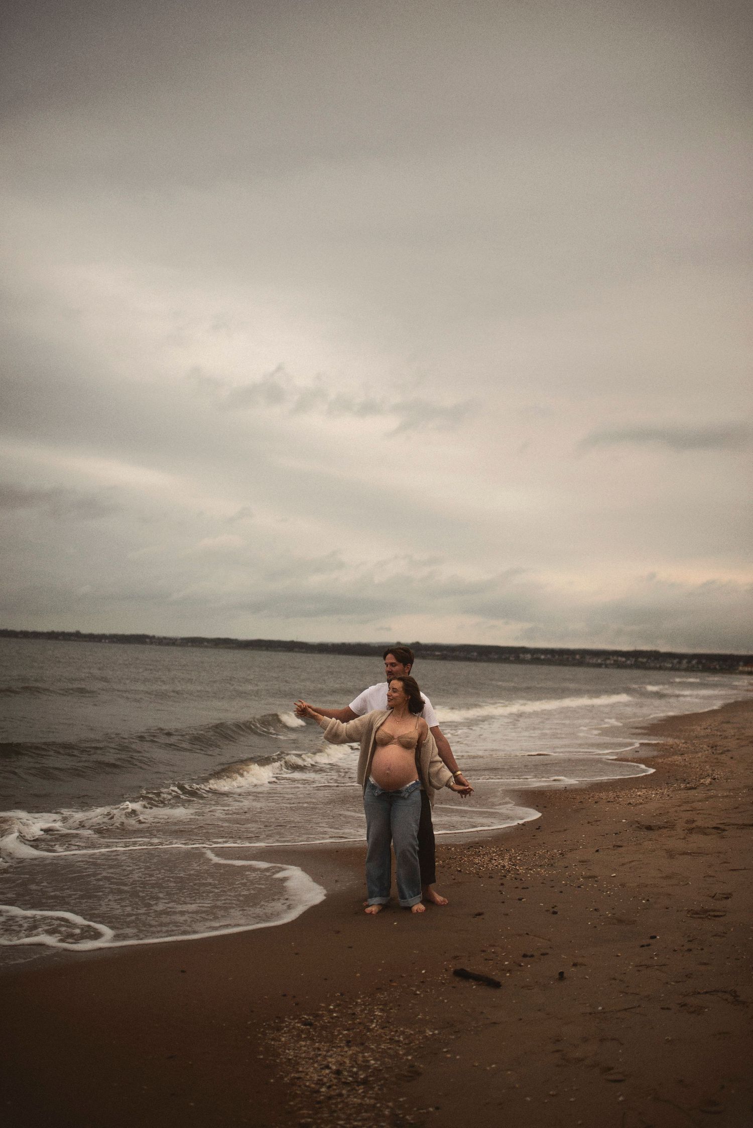 Person with bare feet stands joyfully at the ocean's edge on a cloudy day.