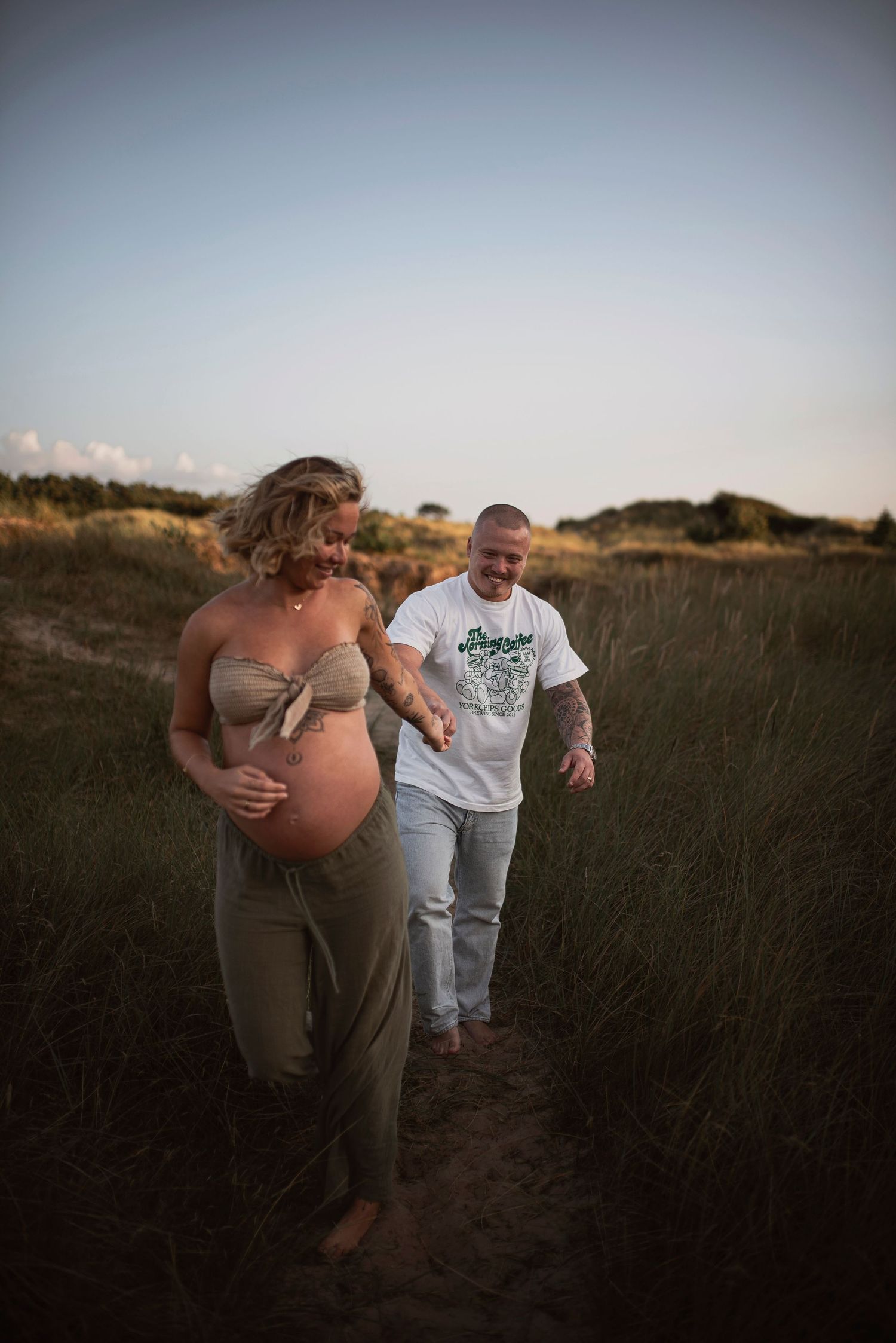 A maternity photoshoot at sunset shows a couple walking together through tall grass in a field.
