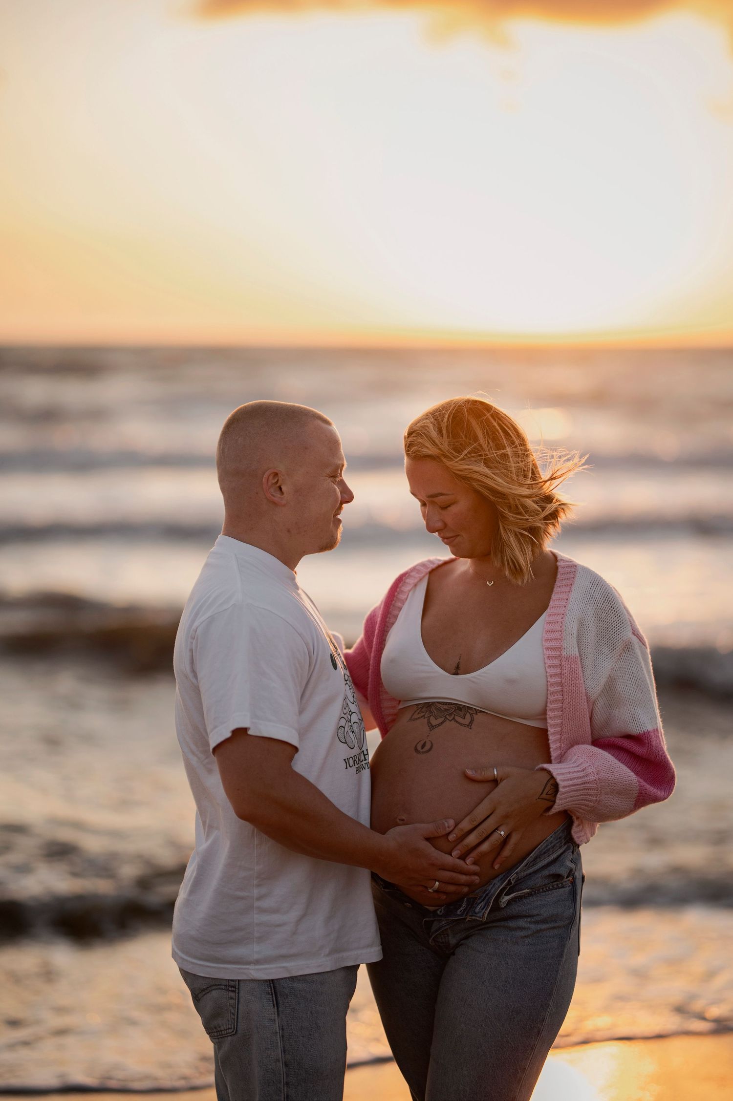 A couple poses for a maternity photo on the beach at sunset with waves and a warm golden sky in the background.