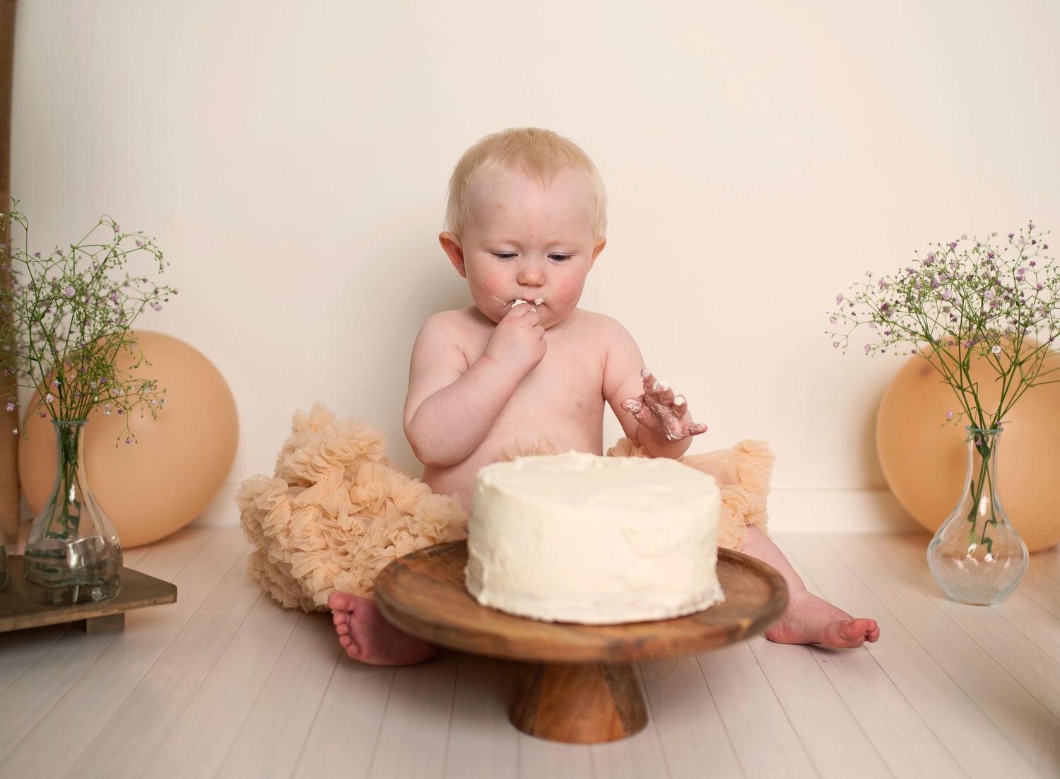 Baby with white cake on wooden stand surrounded by floral decor during cake smash photoshoot.