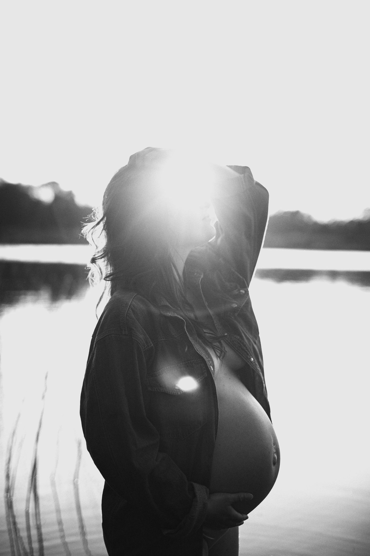 Silhouette of pregnant figure against sunset at a tranquil lake.