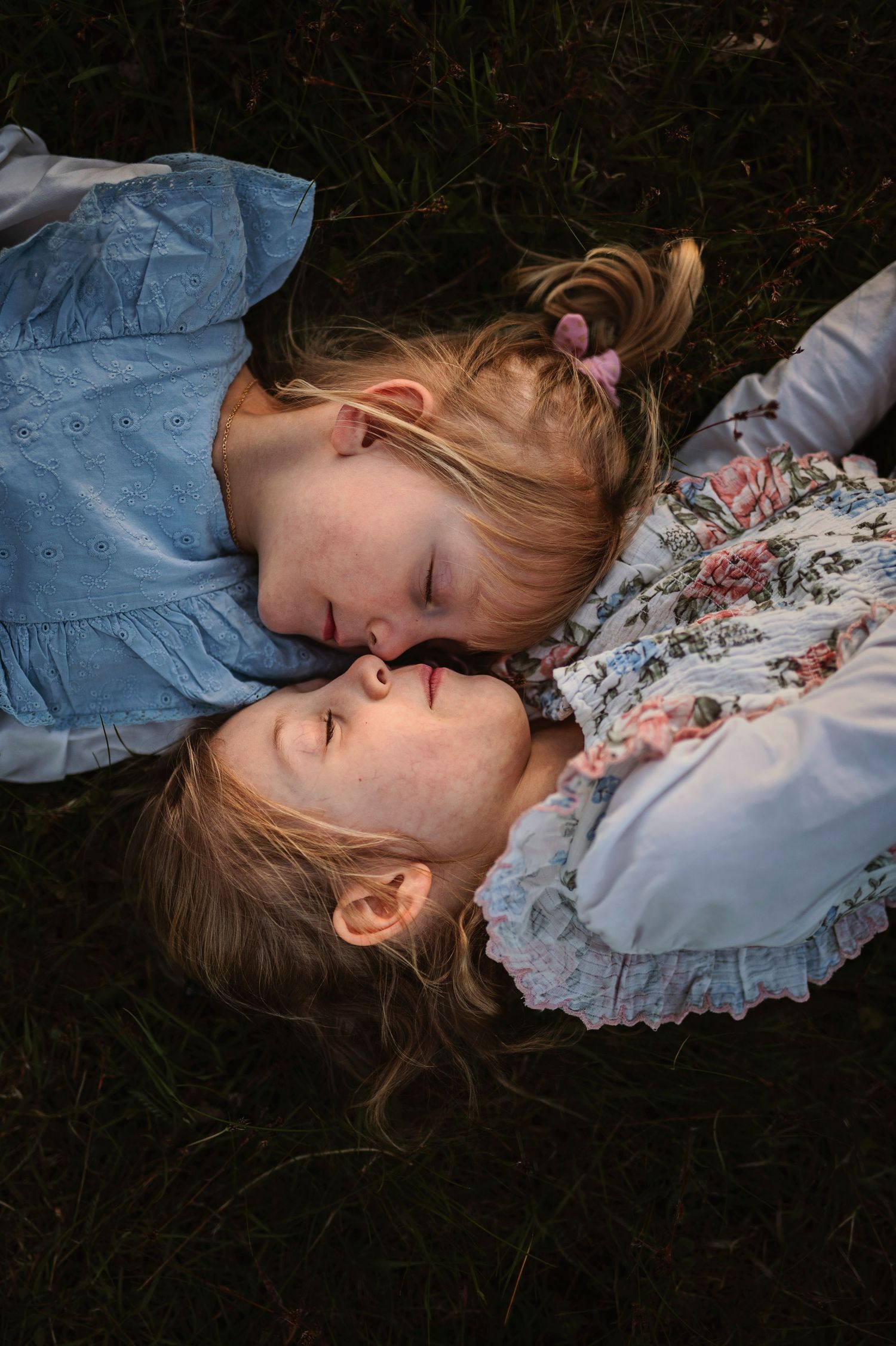Two children lying head to head on grass wearing blue and floral dresses.
