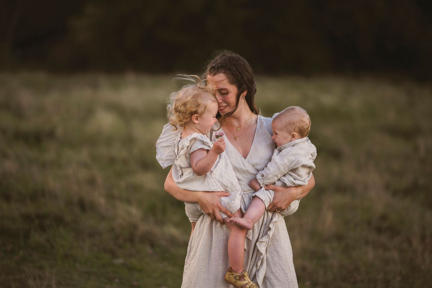 Parent holds two small children while standing in a field at dusk.