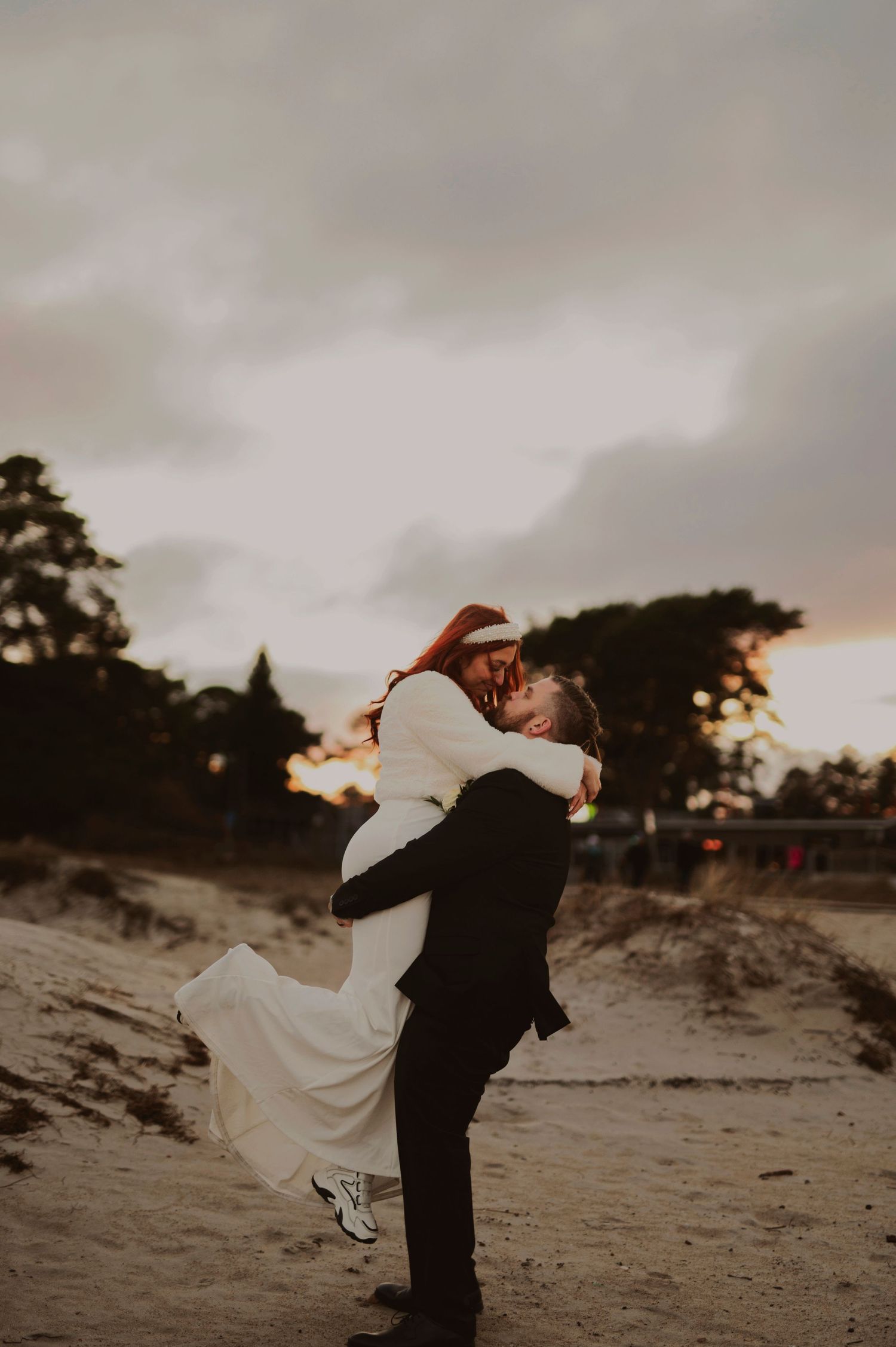 Groom lifts bride in white dress on beach during sunset.
