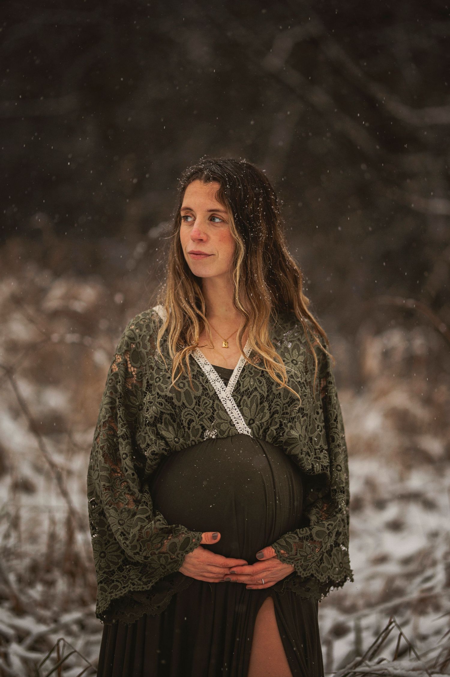 Winter maternity portrait with woman in dark dress and lace cardigan holding belly amidst snowy scenery.