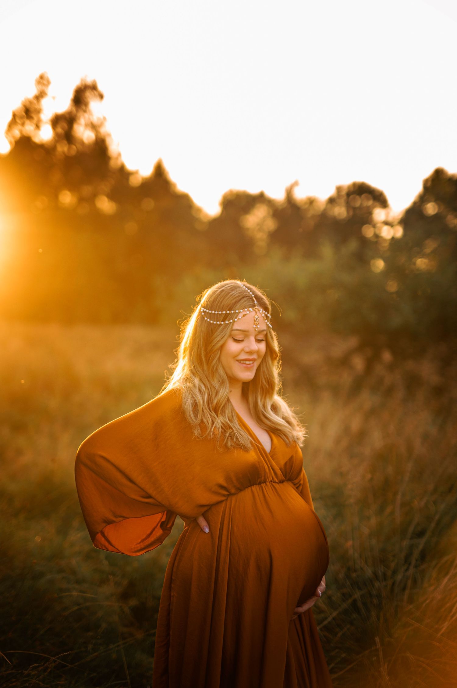 Backlit maternity portrait with flowing rust dress and delicate headpiece during golden hour.