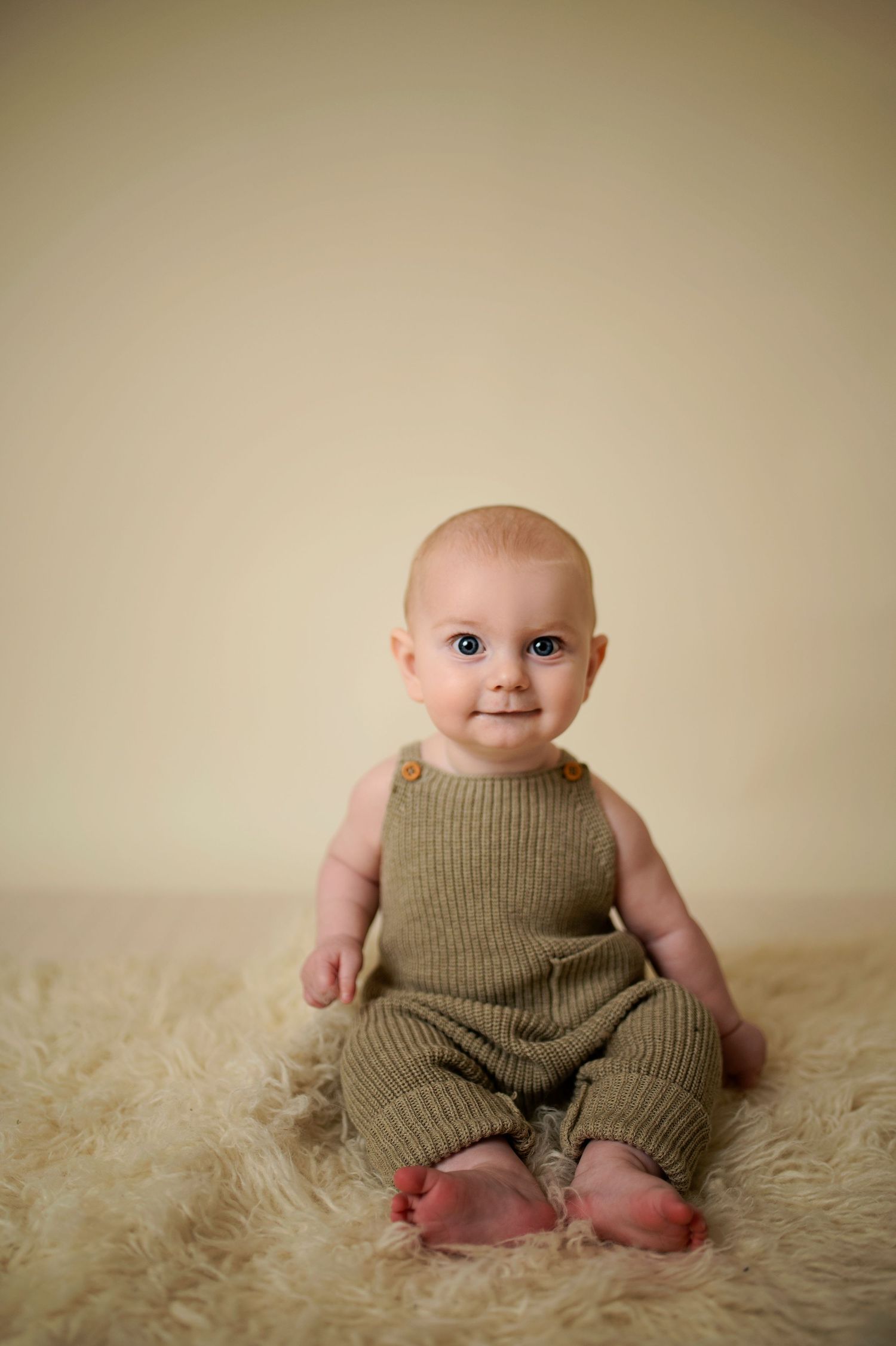 Baby wearing knit romper sits on cream-colored fur rug against neutral backdrop.