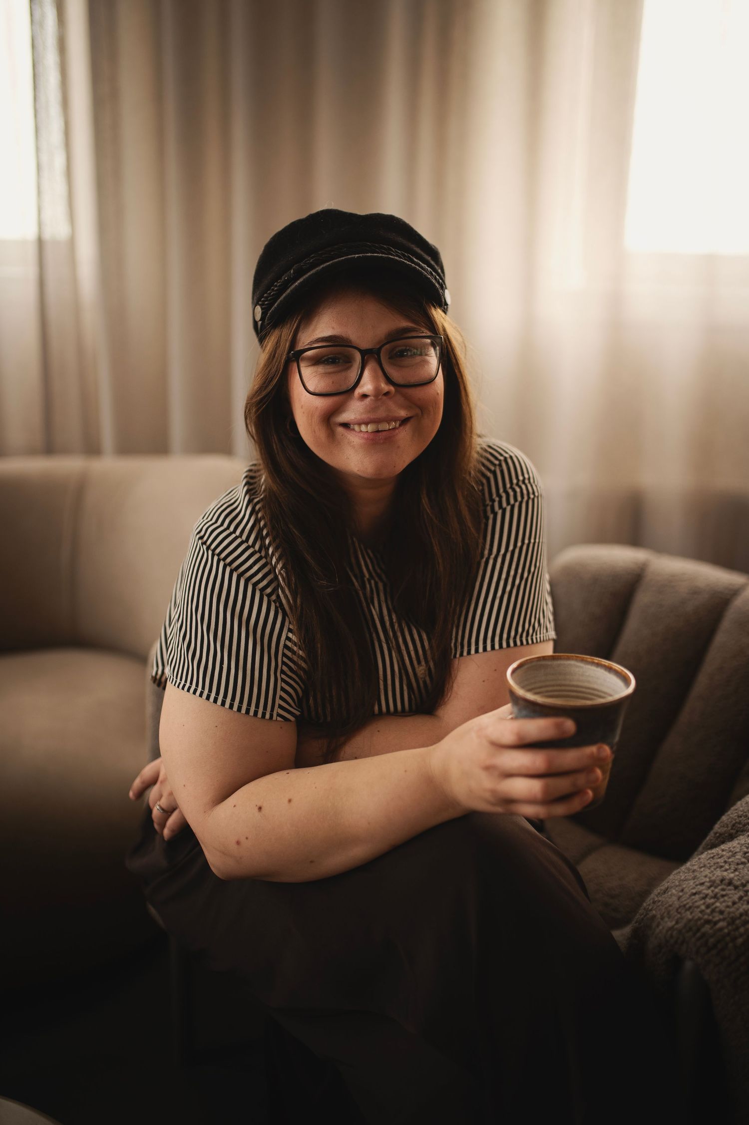 Young woman in striped shirt and black cap smiling on sofa holding a ceramic cup indoors.