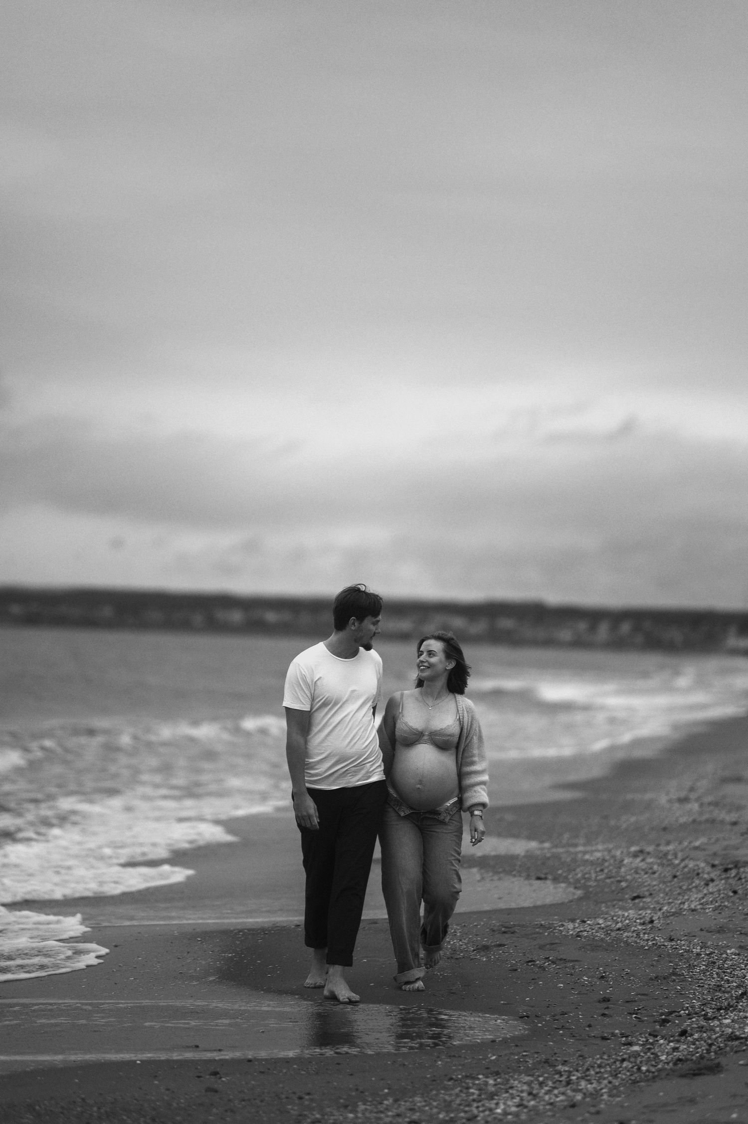 Black and white photo of pregnant couple walking barefoot together on beach shoreline.