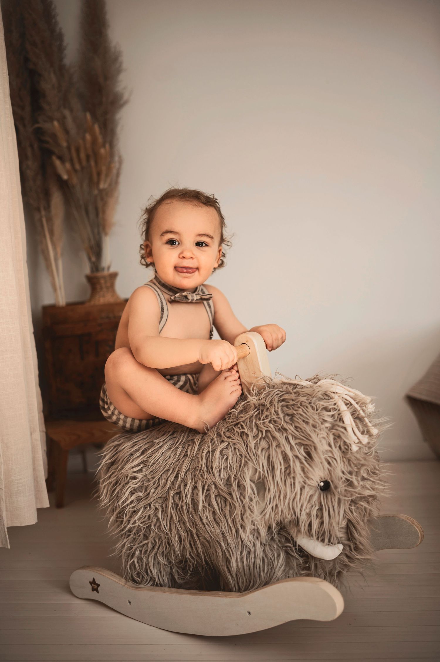 Baby sits on a gray furry rocking toy in neutral-toned nursery setting with pampas grass decor.
