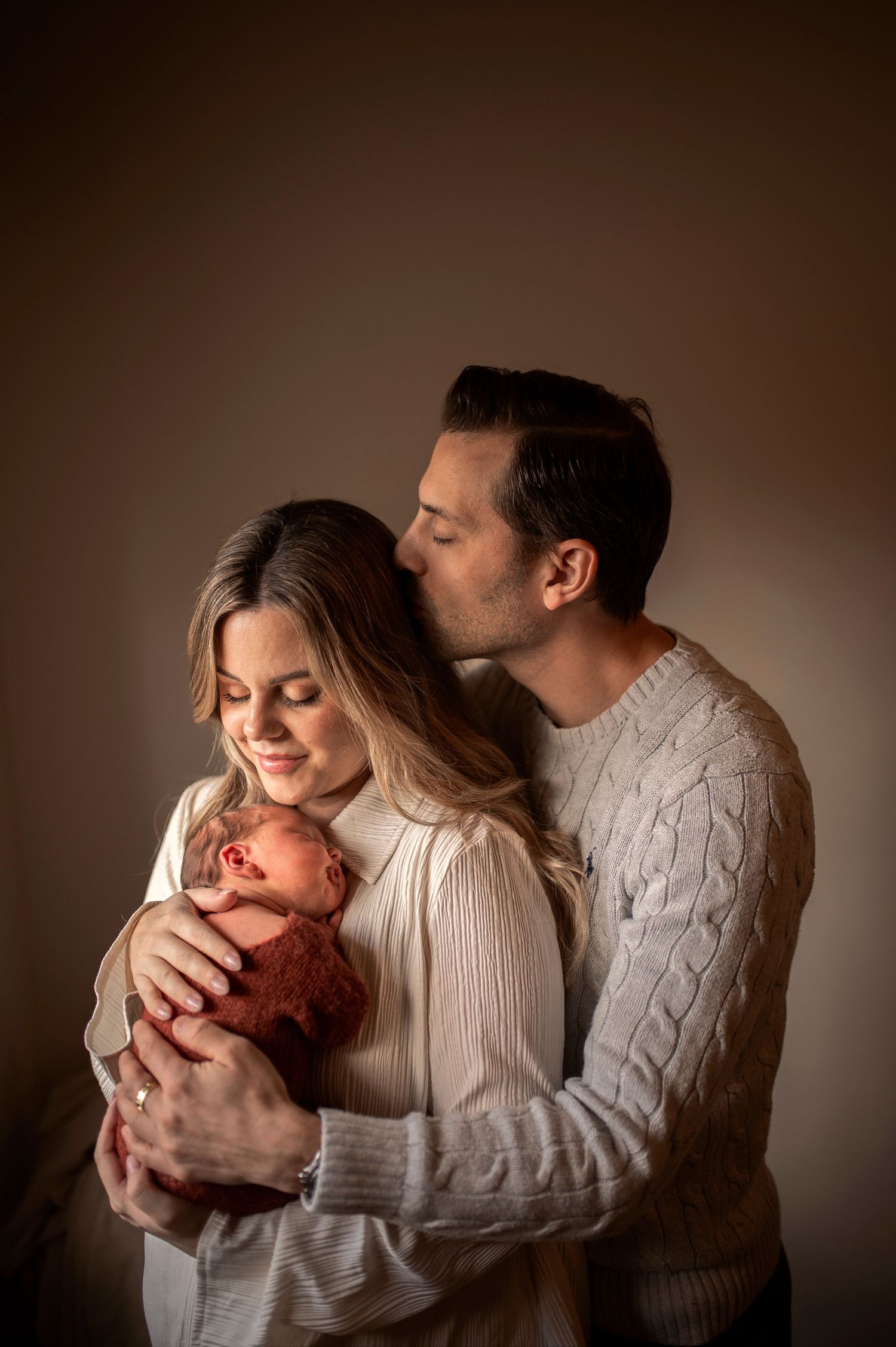 Couple in cozy sweaters embraces their newborn baby in warm studio lighting with brown backdrop.