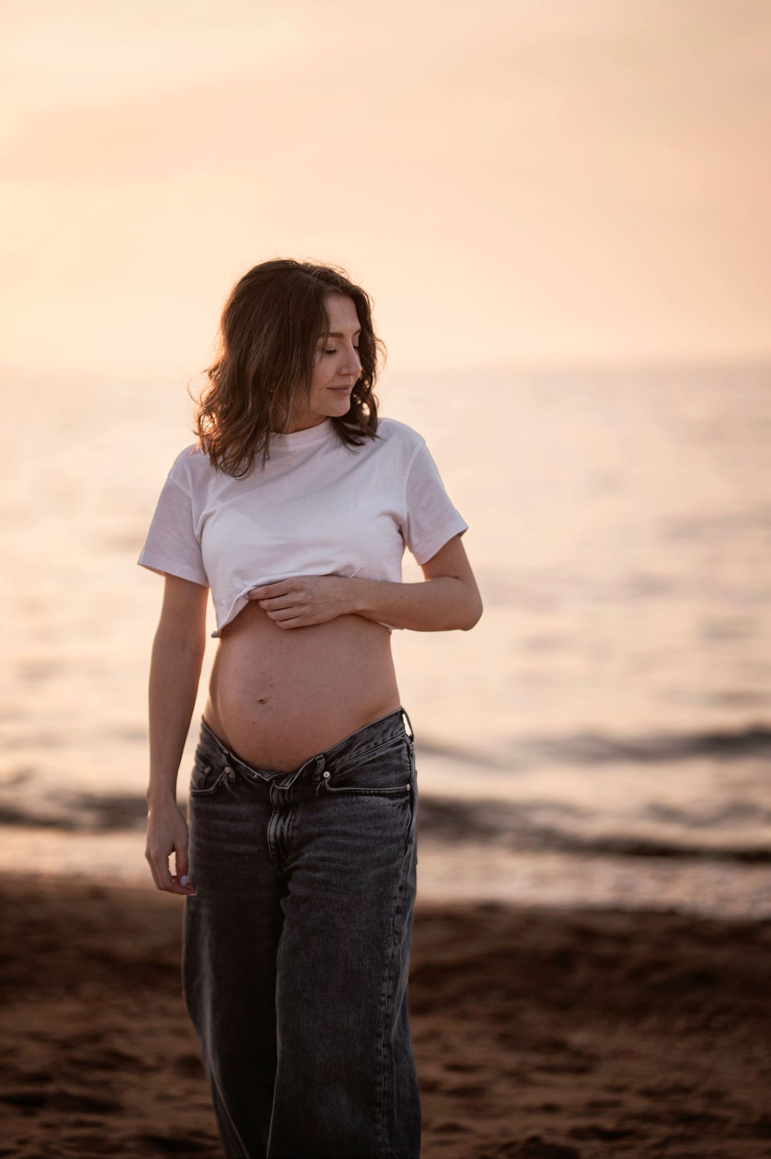 Maternity photoshoot at sunset showing pregnant woman in casual attire on beach.