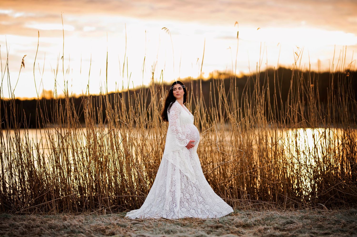 Maternity portrait in white lace dress against golden sunset and lakeside reeds.