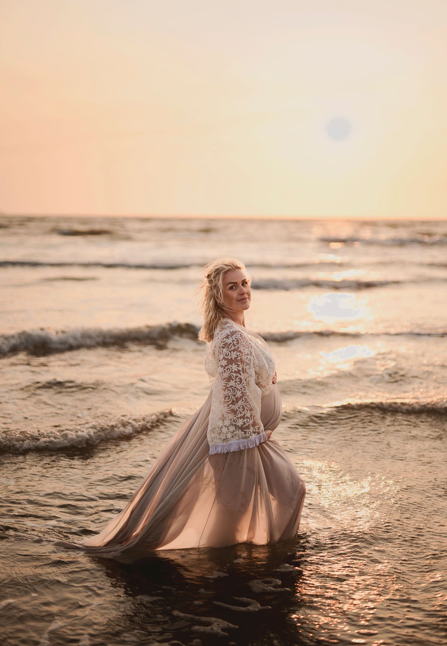 Figure in lace dress stands in ocean waves during golden sunset.