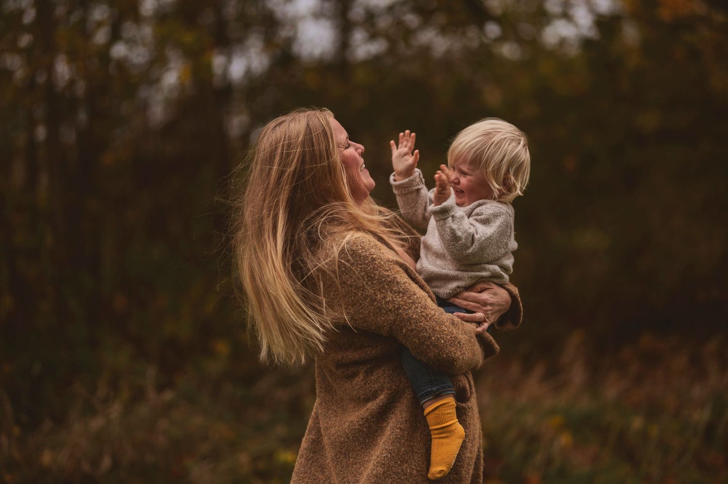 Adult in brown sweater lifts happy child wearing yellow socks against autumn backdrop.