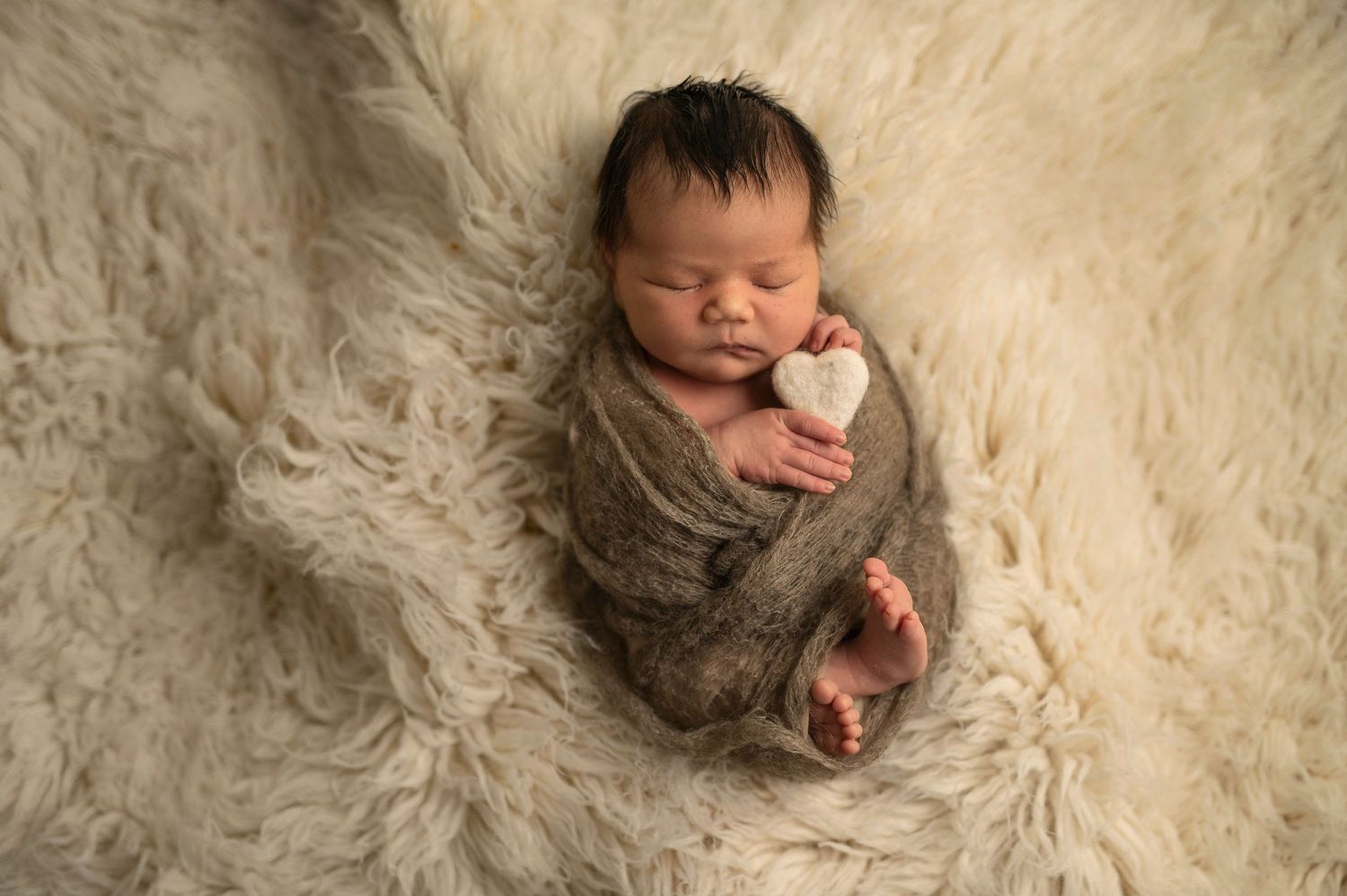 Sleeping newborn wrapped in soft fabric lies on fluffy white fur backdrop.