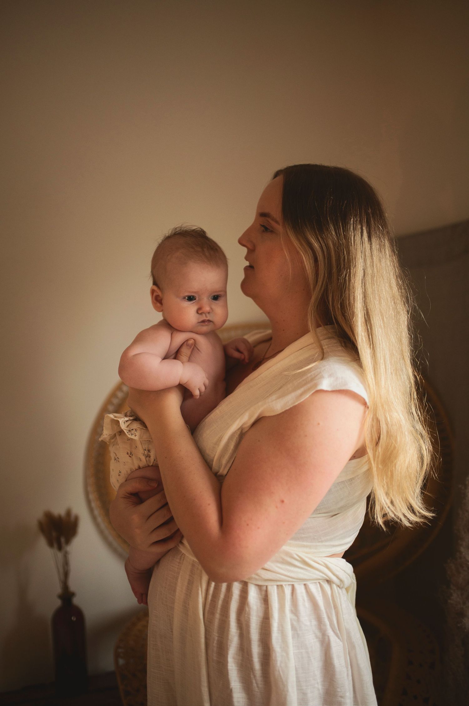 Mother in white dress holding infant in warm-toned indoor setting.