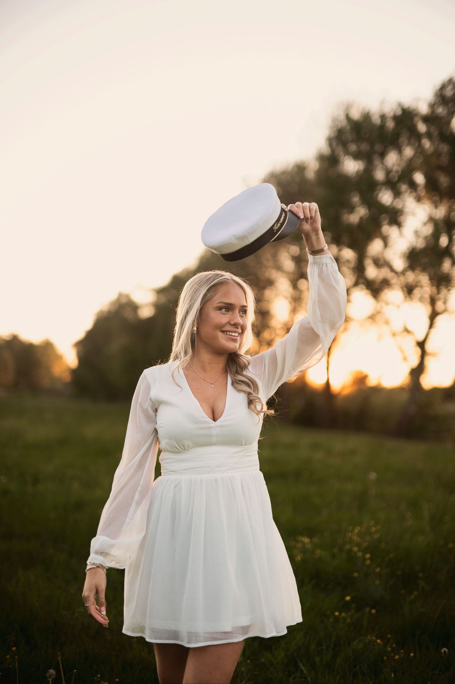 Smiling blonde woman in white dress holding captain hat up in a sunset meadow.