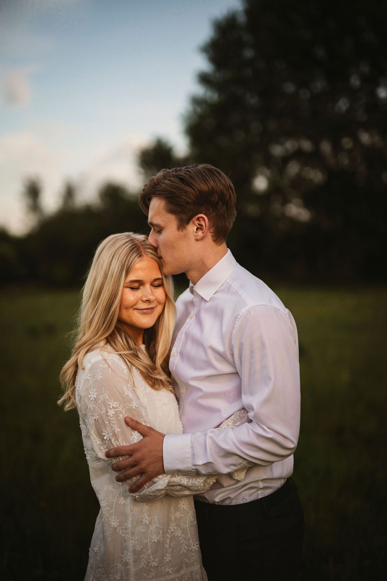 A couple embraces at golden hour in a field, the man kissing the woman's head as she smiles in a lace dress.