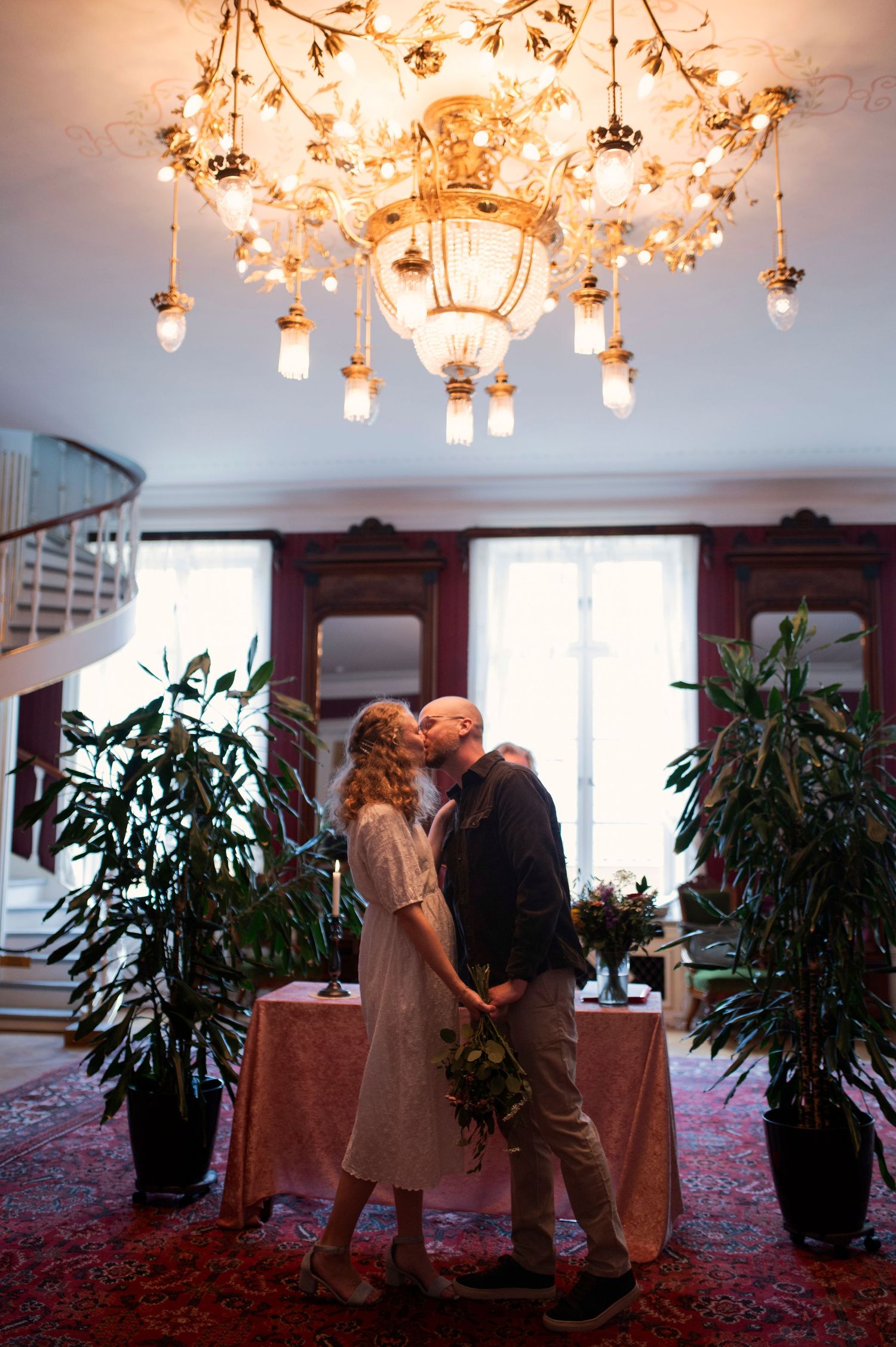 Couple sharing a romantic kiss under an ornate golden chandelier in an elegant venue with lush plants.