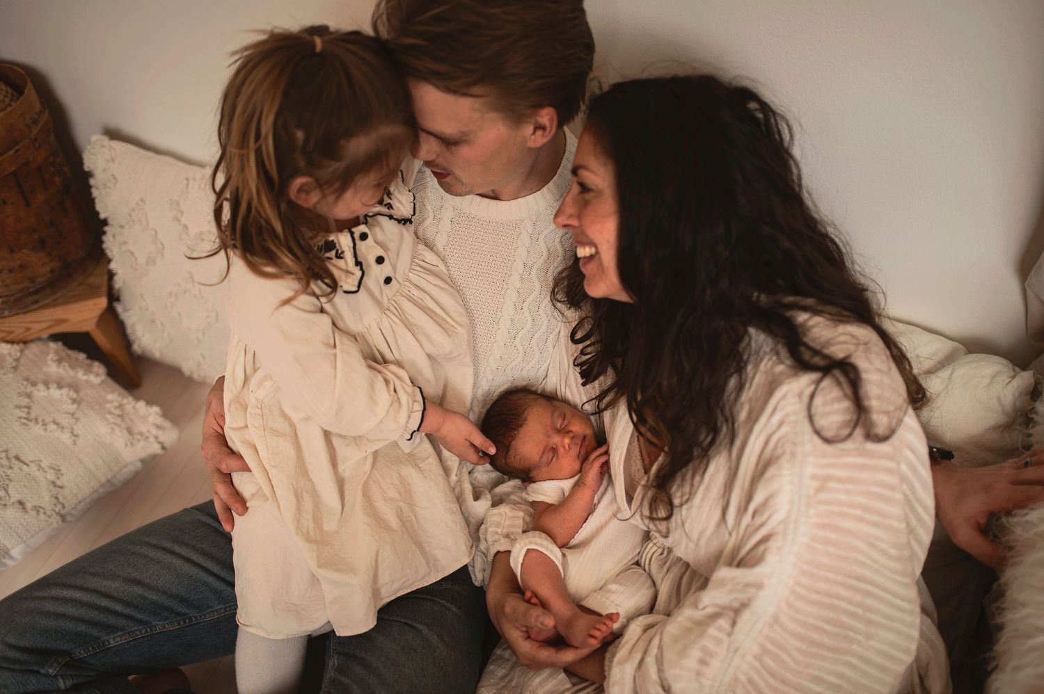Family of three with toddler girl admiring newborn baby on white bed.