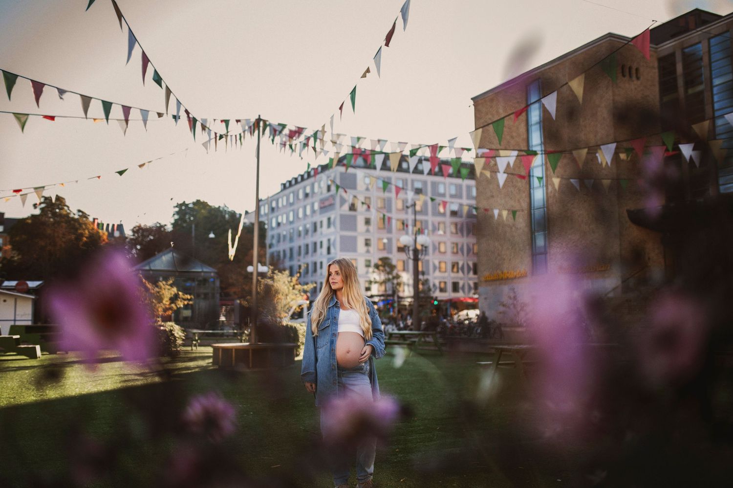 Pregnant woman standing in a sunny park with colorful bunting flags and pink flowers.