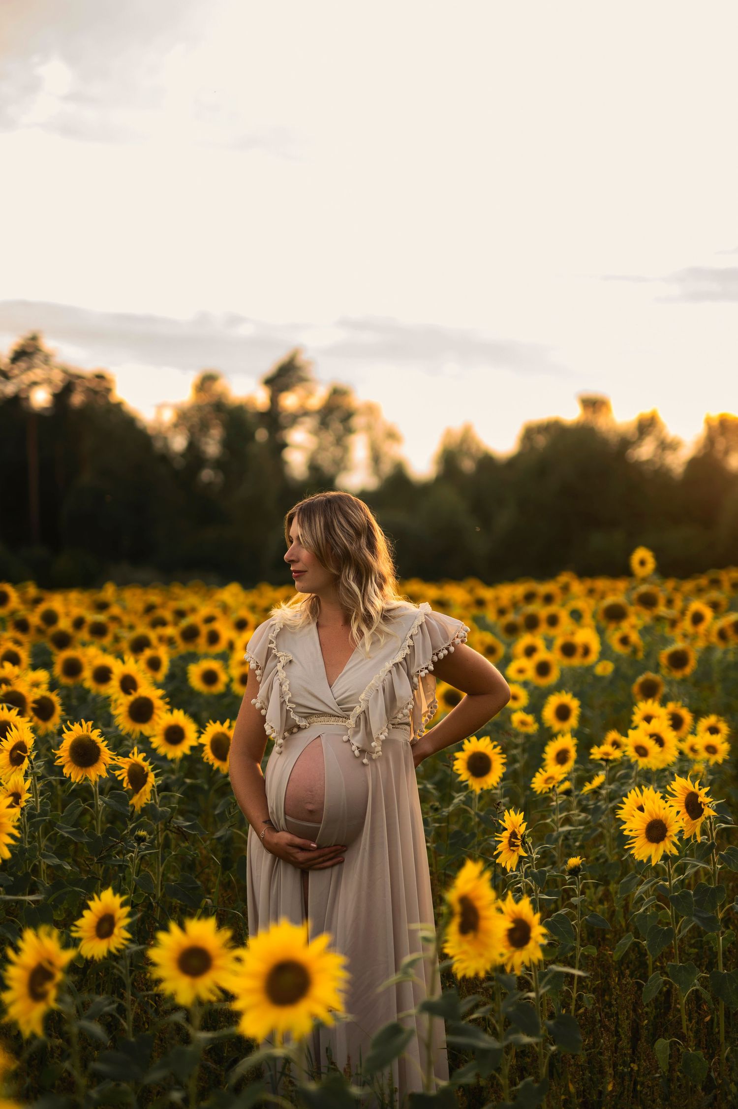 Maternity portrait in flowing dress amidst golden sunflower field at dusk.