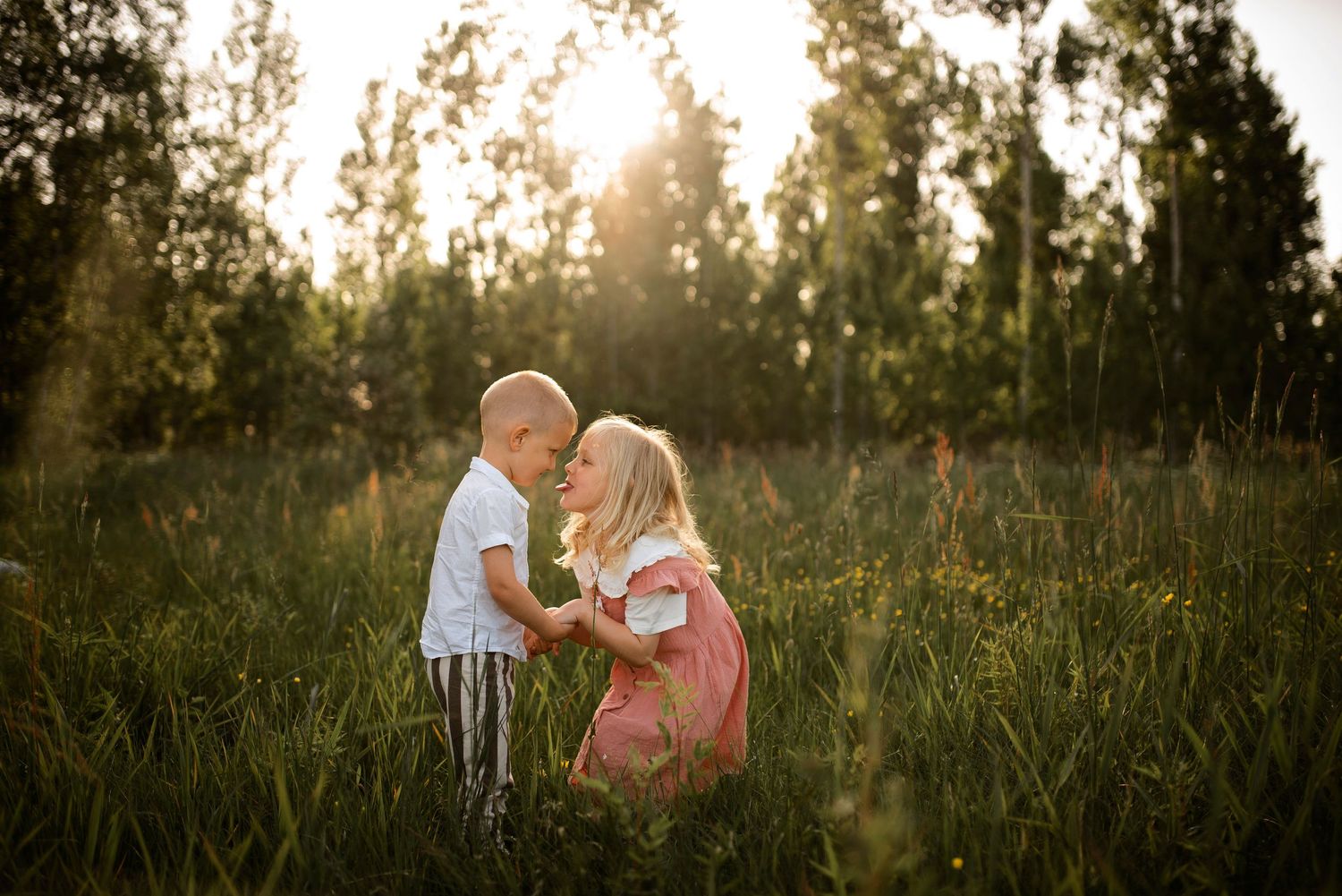 Two children playing in tall grass with sunlit trees in background.