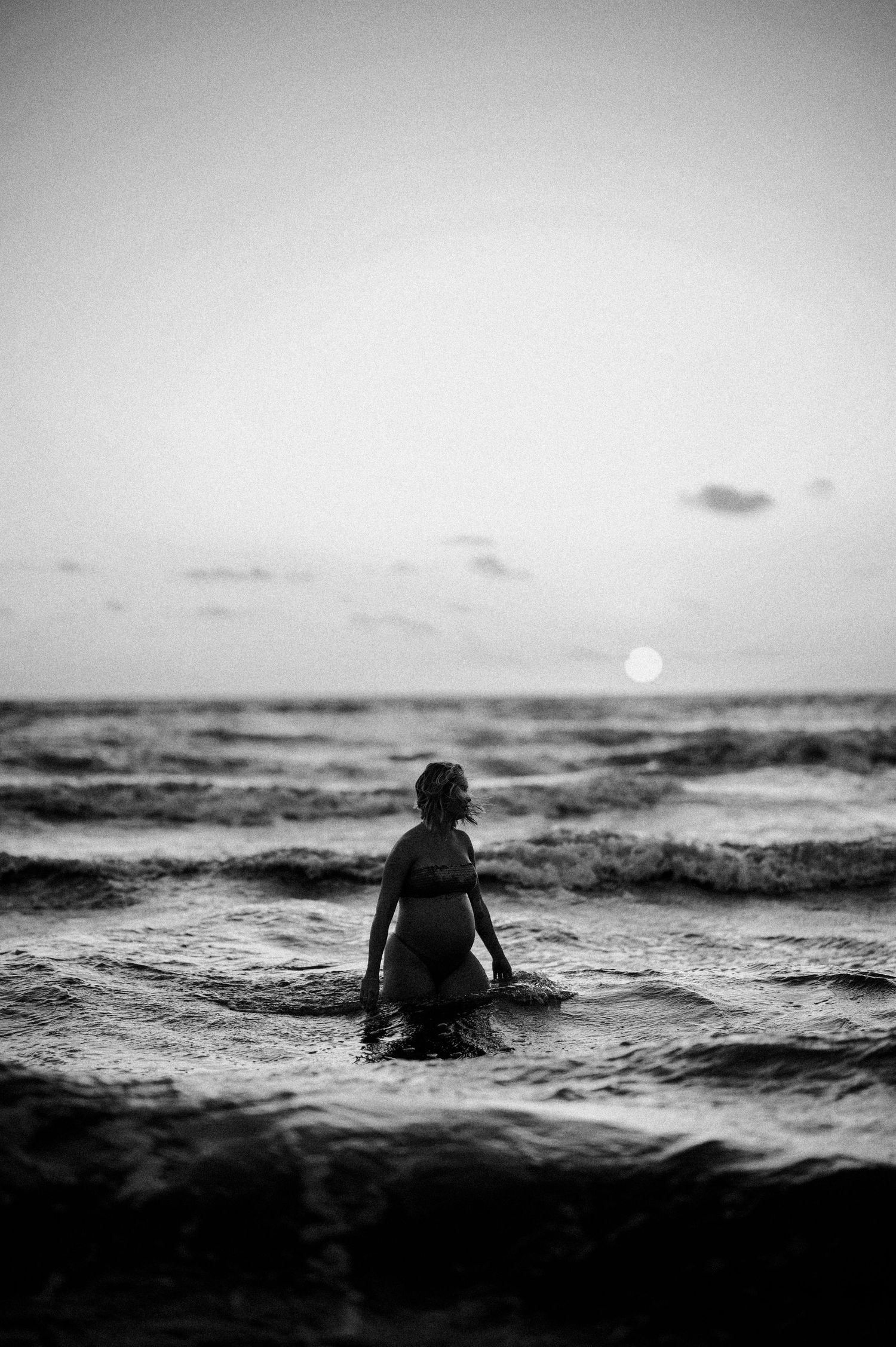 A silhouetted figure swims in dark ocean waves at sunset in dramatic black and white.
