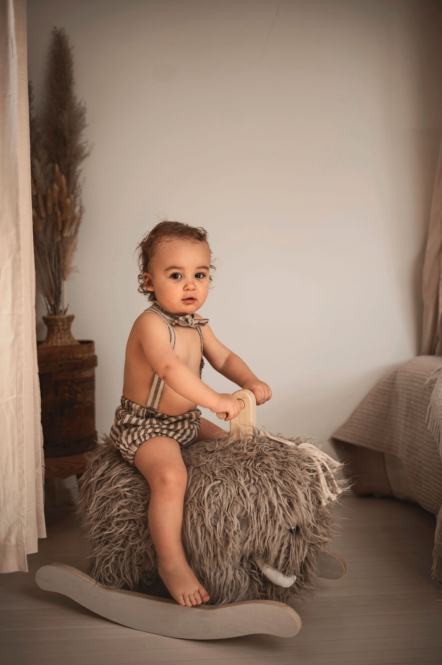Young child wearing striped outfit sits on fluffy gray rocking horse in neutral-toned studio setting.