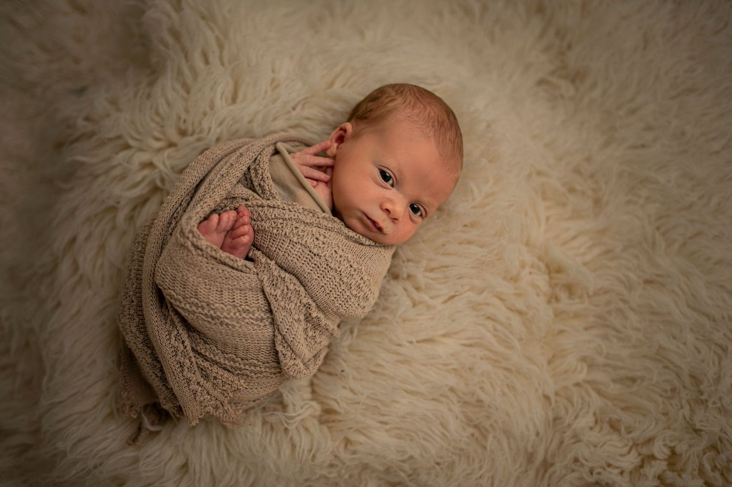 Newborn baby wrapped in beige knit blanket lying on soft white furry backdrop.
