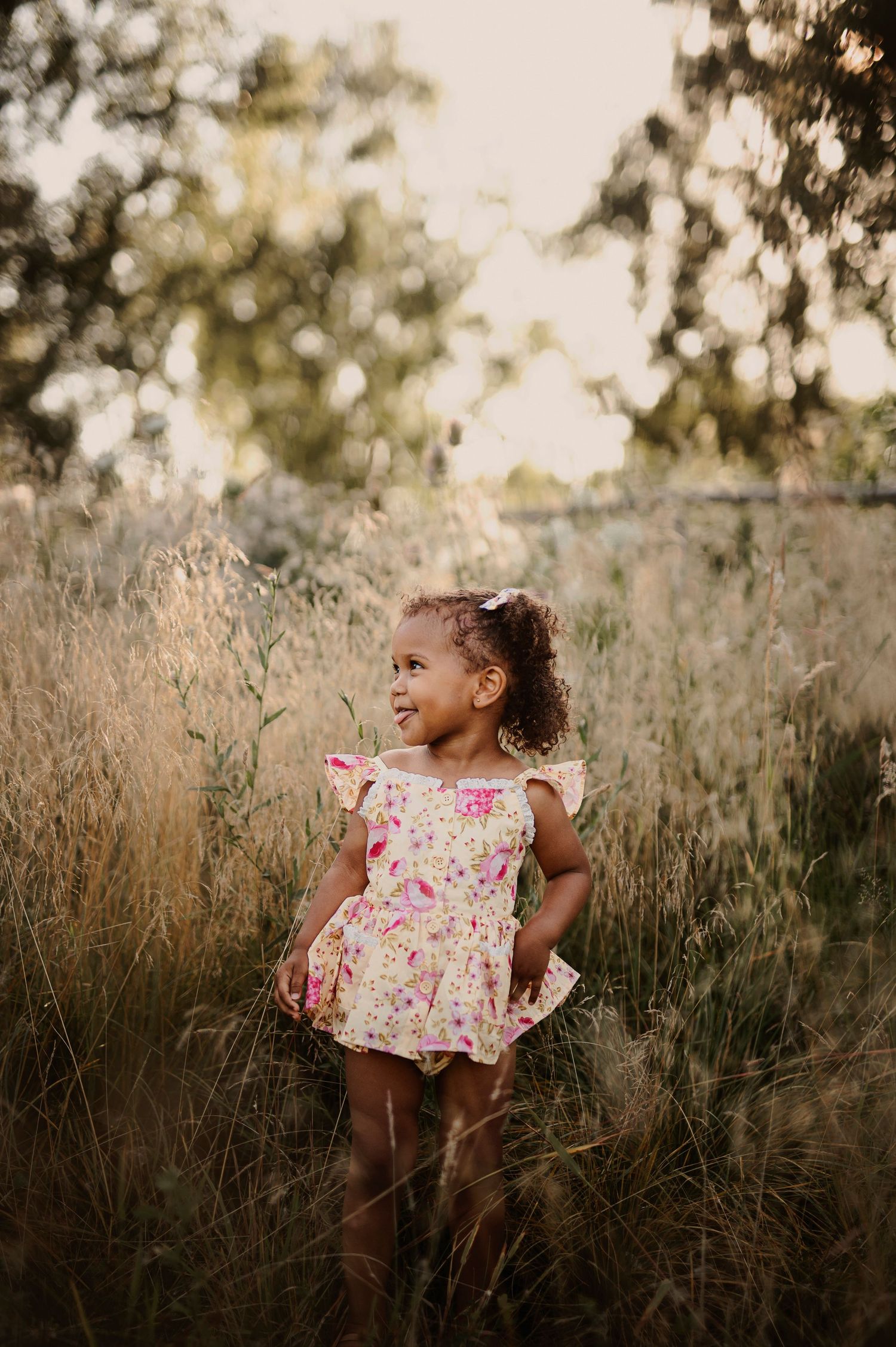 A joyful toddler in a floral dress stands in a sunlit field of tall grass during golden hour.