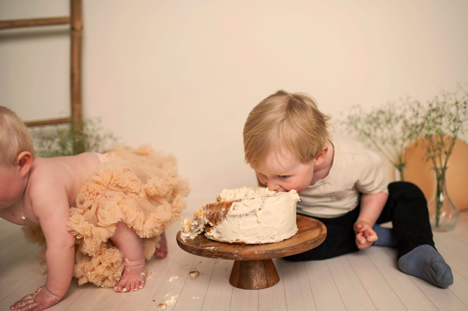 Two babies enjoying messy cake smash session with white frosted cake on wooden stand.