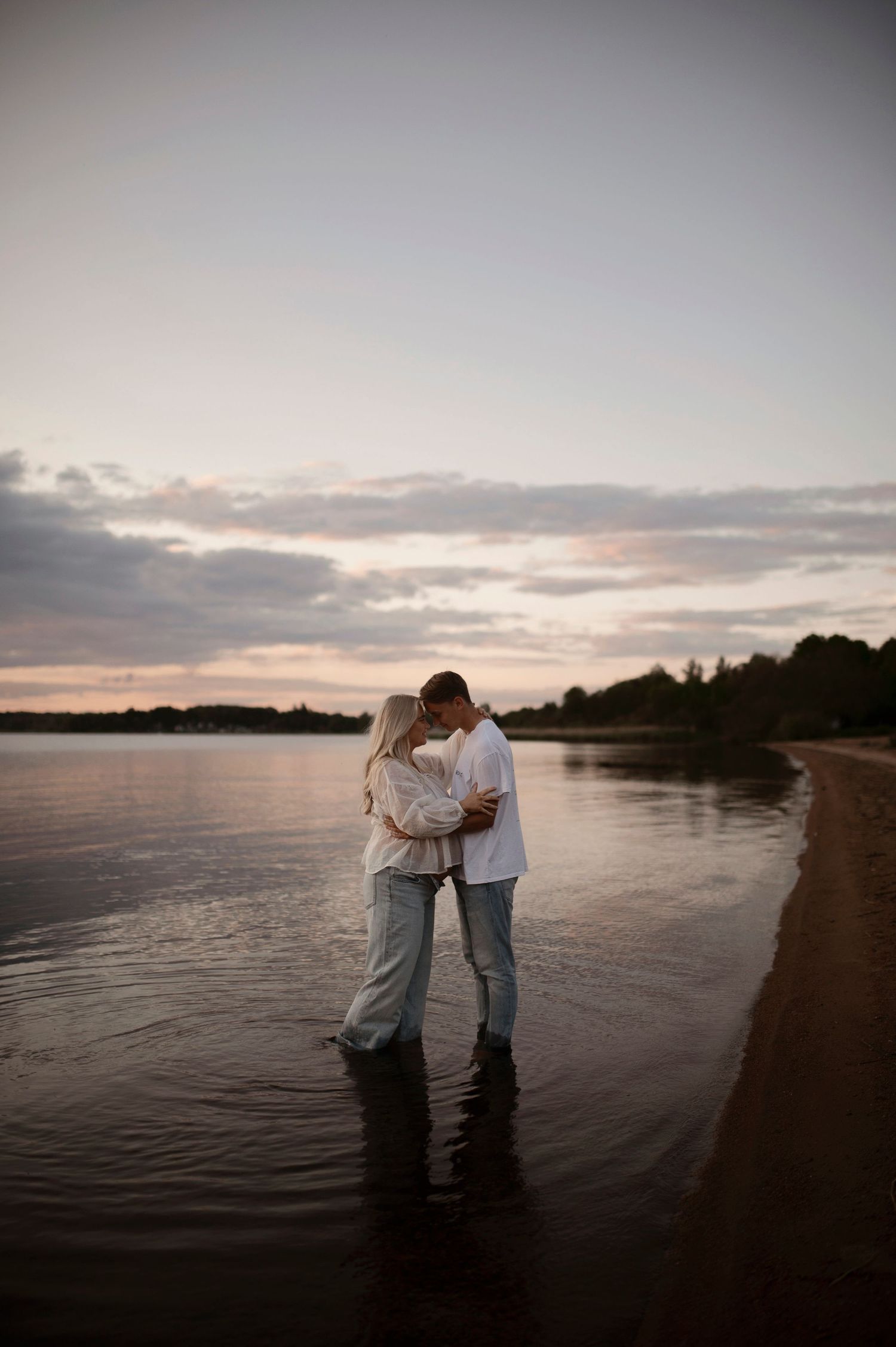 A couple stands embracing in shallow water at sunset with a dramatic cloudy sky backdrop.