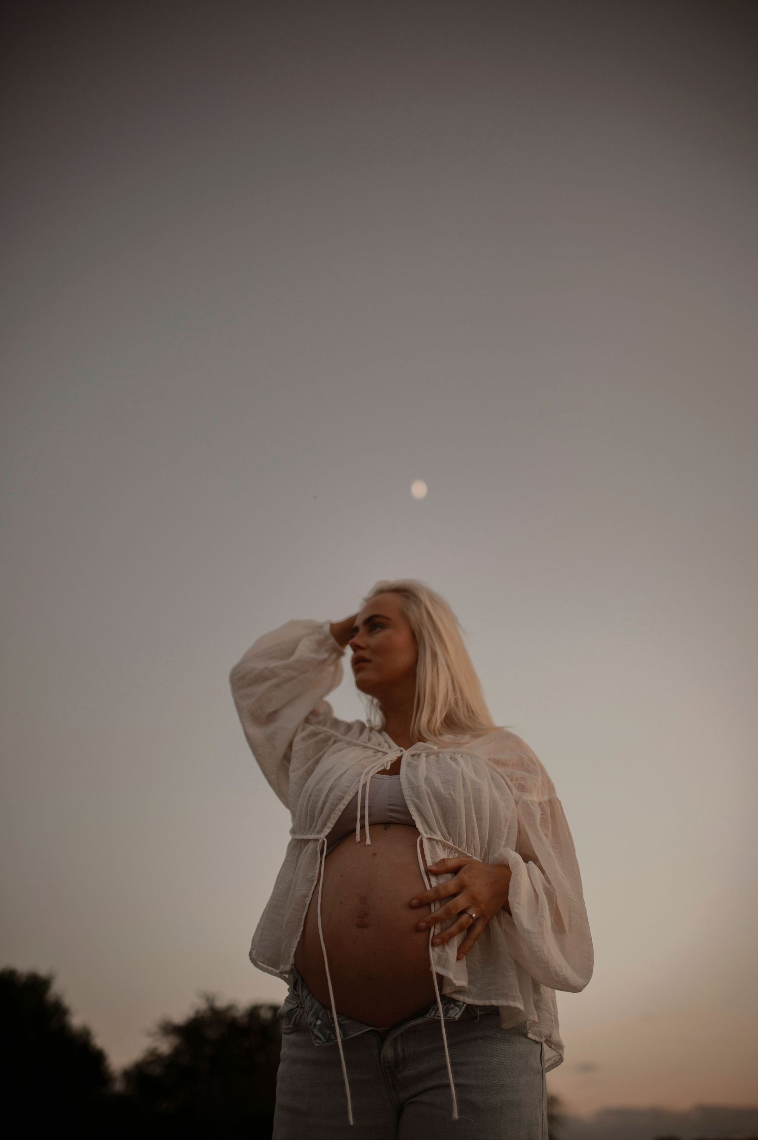A silhouette poses in a white peasant top during sunset with the moon visible in the hazy sky.