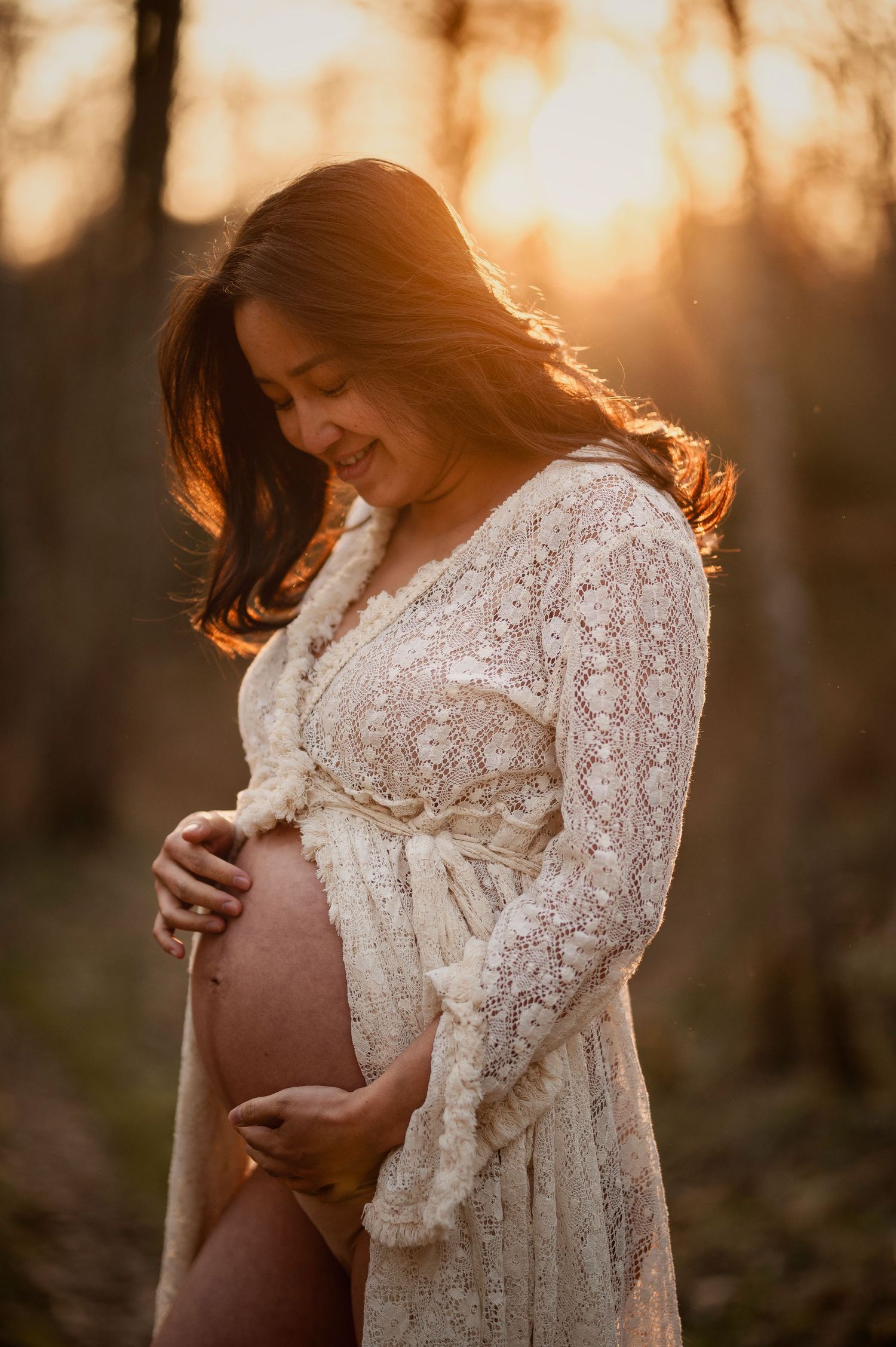 A pregnant woman in a white lace dress stands in golden sunlight during sunset outdoors.