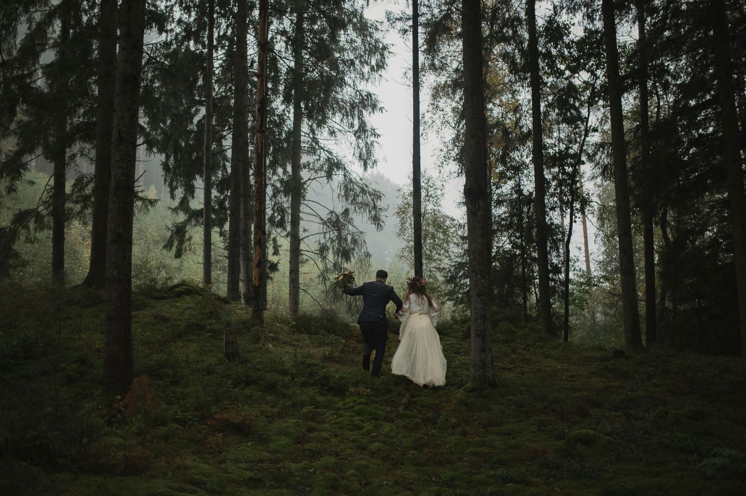Bride and groom walking hand in hand through misty green forest during wedding.