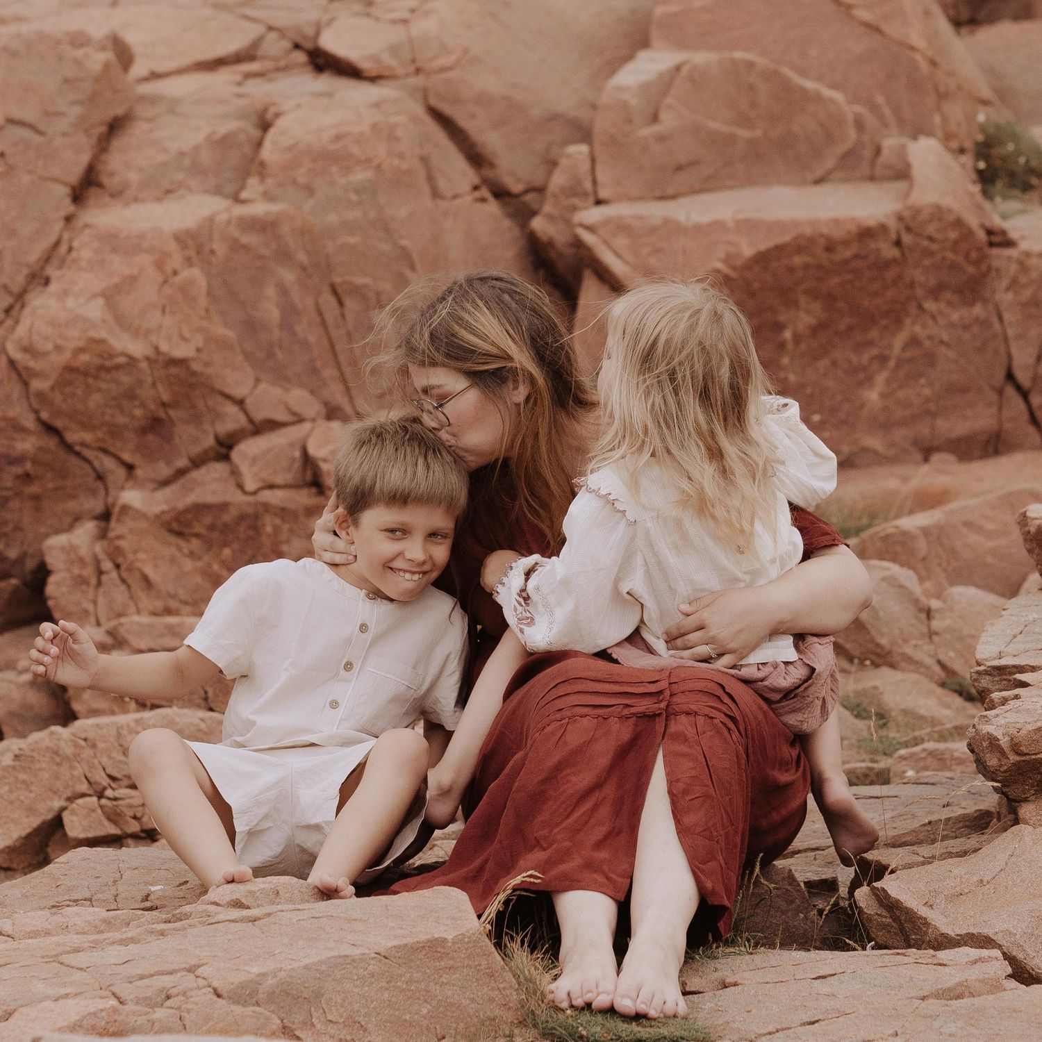 A mother sits on red rocks with her two young children, sharing a tender moment outdoors.