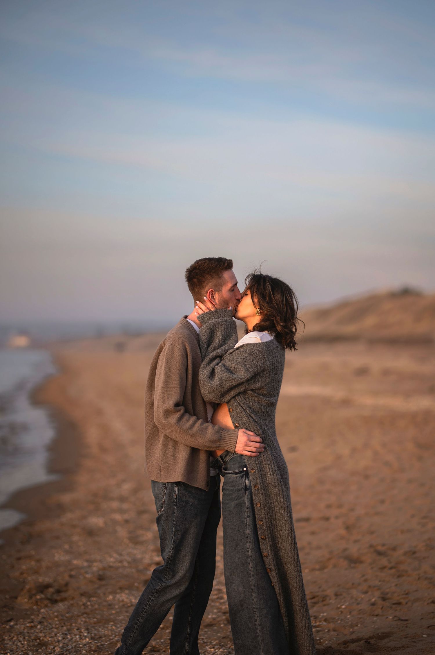 A couple shares a romantic kiss on a peaceful beach at dusk while wearing cozy sweaters.